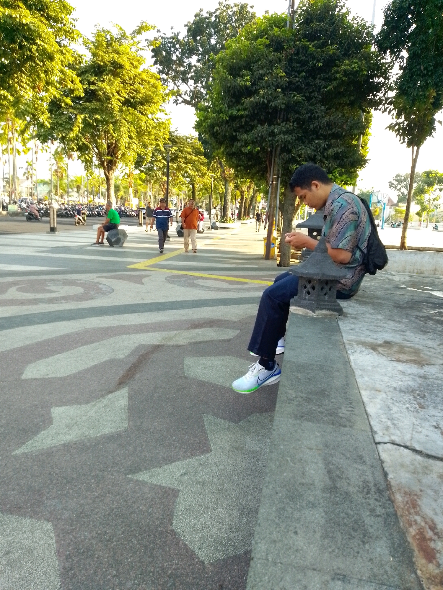 The image depicts an outdoor scene in the city of Jember, Indonesia, likely a pedestrian area adjacent to a street. In the foreground, a young man is seated on a concrete ledge, looking down at his phone. He is wearing a patterned short-sleeved shirt, dark trousers, and white athletic shoes with blue and green accents. A black backpack is slung over his right shoulder. Beside him on the ledge is a small, dark, decorative lantern. The ground in front of the ledge is paved with asphalt and features a geometric pattern of interlocking shapes in shades of gray and white.
In the mid-ground, several people are walking. One man in a green shirt and dark shorts sits on a large, rough-textured stone. Further back, two men are walking towards the left of the frame; one wears a blue and white striped shirt and dark pants, while the other wears an orange shirt and light-colored pants. Behind them, a child in a blue shirt and dark pants walks, and an adult in an orange shirt is visible. To the left of the pedestrian area, a row of parked motorcycles is visible, suggesting proximity to a road.
The background is dominated by lush green trees, including tall trees with bright green foliage and denser, darker trees. Sunlight filters through the leaves, creating a dappled effect. Streetlights are also present, lining the pedestrian walkway. The overall impression is a public space in Jember, likely a park or promenade, on a sunny day.