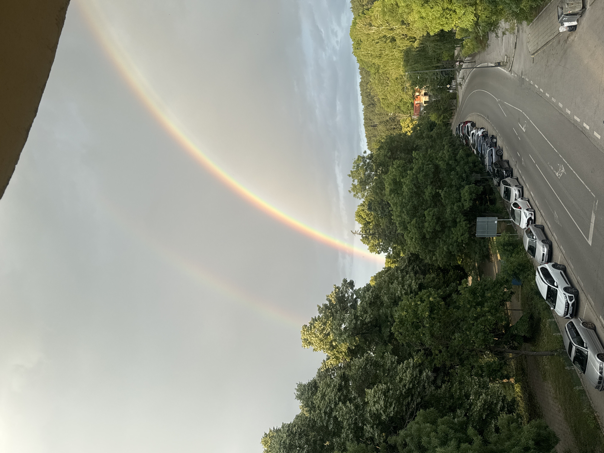 This image depicts a view of a street in Bankya, Bulgaria, with a double rainbow visible in the sky. The street is lined with parked cars and mature trees. The sky is overcast with clouds, creating a diffused light. In the foreground, there is a section of a building's facade visible at the top edge of the frame. The road curves to the right, leading into a forested area with a few buildings interspersed amongst the trees. Several cars are parked along the side of the road and in a designated parking area. The overall scene suggests a natural setting within a residential area.