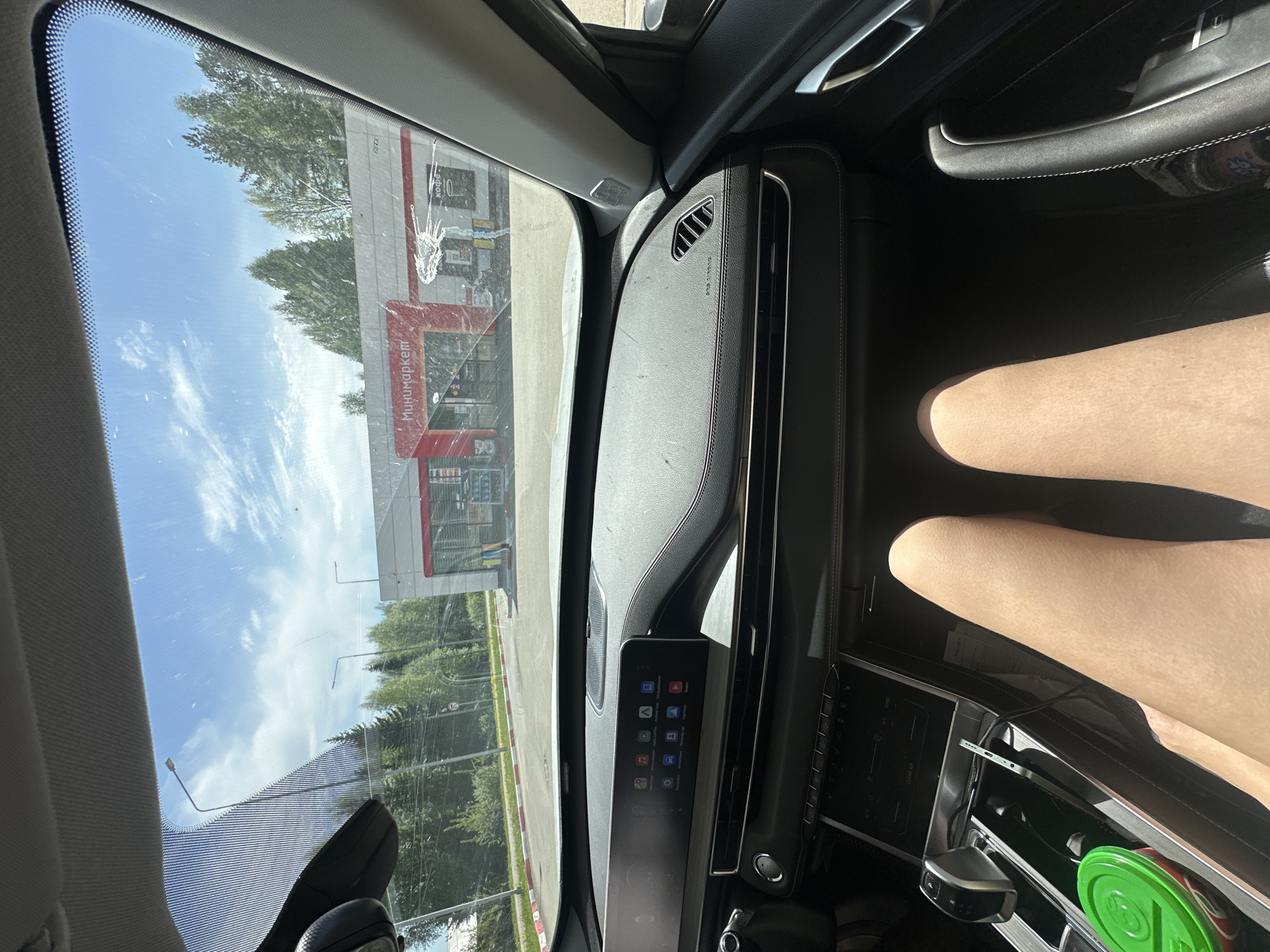 The image shows an interior view from inside a car, looking out the windshield toward a minimarket. The market is a single-story building with a red roof and signage in Russian. Outside, there are trees and a parking area. Inside the car, a dashboard with a screen displaying app icons is visible. A person's legs are visible in the lower right of the frame. A green cup is in the center console.
The location is Uva, Russia.