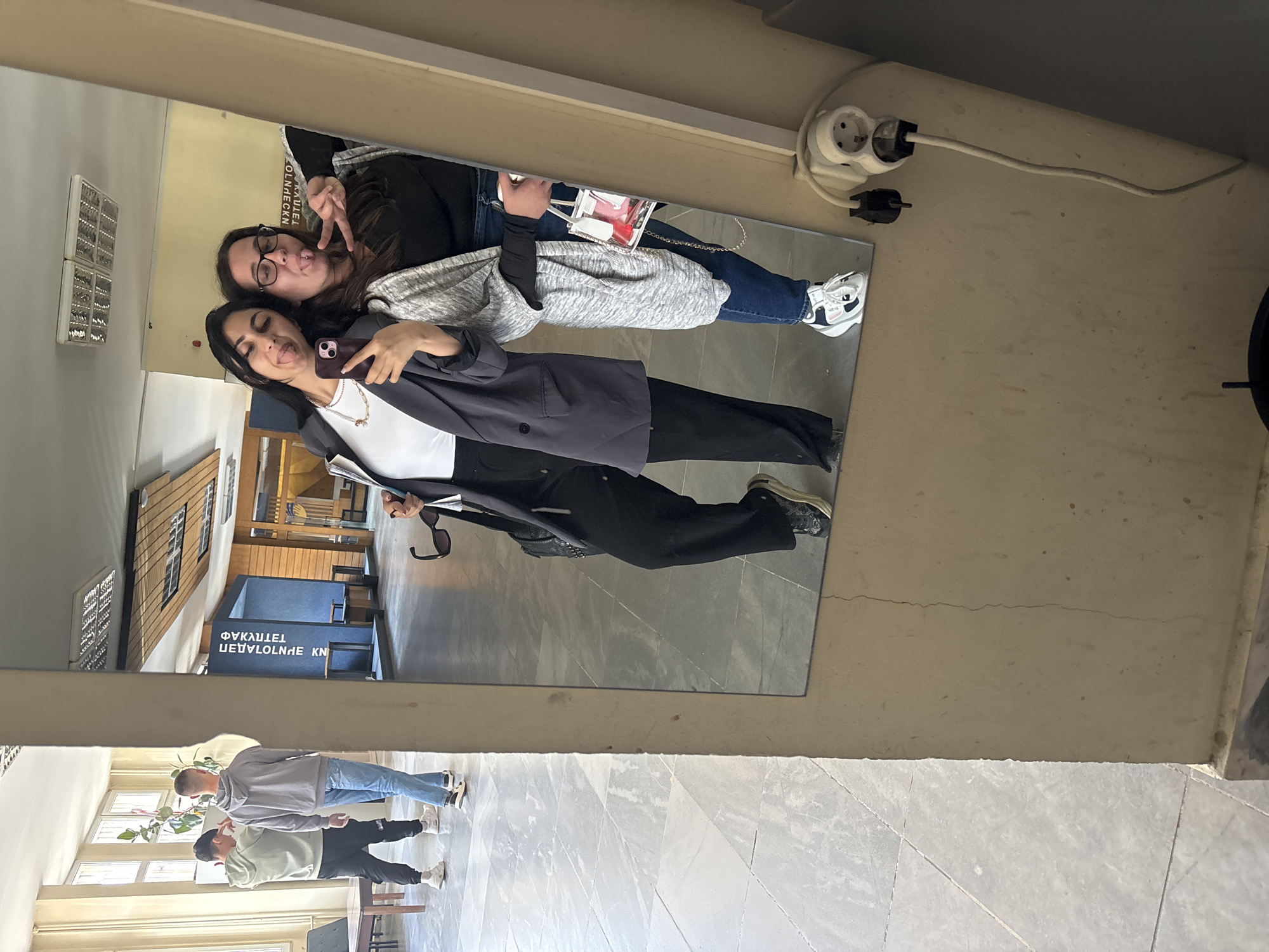 Two women are taking a selfie in a mirror. One is wearing a black blazer, black pants, and a white top with a necklace, and holding a phone. The other is wearing jeans, a black shirt, a gray cardigan, and glasses. The reflection in the mirror includes other people and text "ФАКУЛТЕТ ЛОЛИНЕСКN". An electrical outlet and plug are visible above the mirror. The mirror is in a hallway with tile flooring and some visible people. This hallway could be in the Faculty of Technology at the University of Food Technologies in Plovdiv, Bulgaria, judging by the mirrored text.