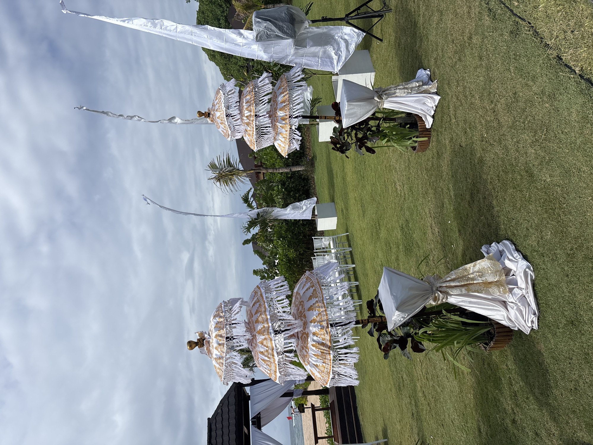 The image depicts an outdoor wedding or event setup on a grassy lawn overlooking a beach. In the foreground, there are two ornate, tiered ceremonial umbrellas with white and gold fabric and fringes, placed on stands. Each stand is draped with white and gold fabric. Several tall, white decorative flags are also visible, fluttering in the breeze. In the middle ground, rows of white chairs are arranged facing the ocean, and a gazebo with white draping stands near the beach. The background shows lush green foliage and palm trees. The sky is overcast with clouds. The scene suggests a preparation for an event in a tropical coastal location in Kuta Selatan, Indonesia.