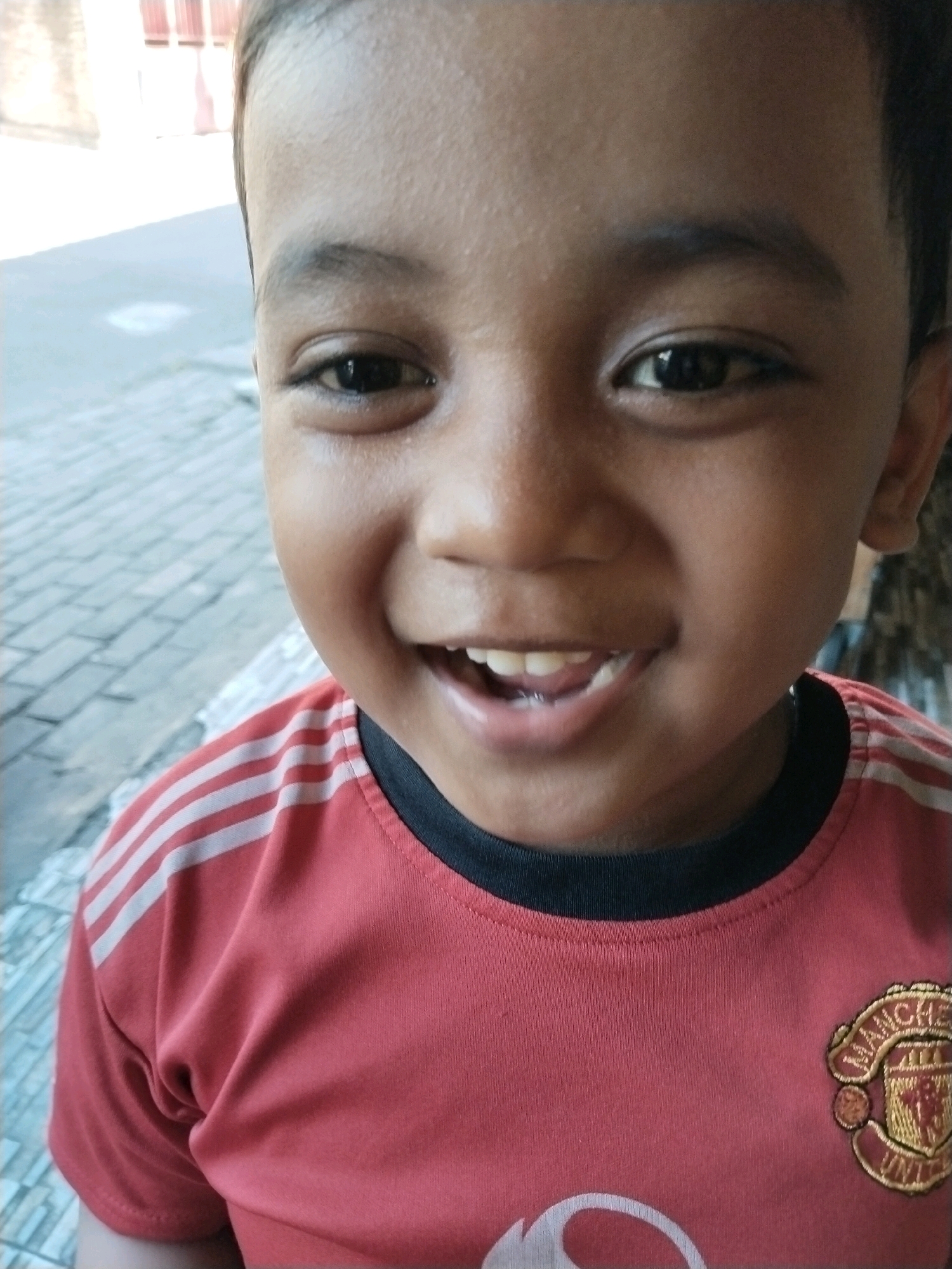 The image is a close-up, waist-up photograph of a young boy with brown skin, dark eyes, and dark hair, smiling directly at the camera. He is wearing a red t-shirt with white stripes on the sleeves and a black collar. A Manchester United logo is visible on the right side of his chest. The background is blurred but appears to be an outdoor setting with a paved surface made of interlocking bricks and a building with a red roof in the distance. The lighting suggests it is daytime. The photograph was taken in Pepelegi, Indonesia.
