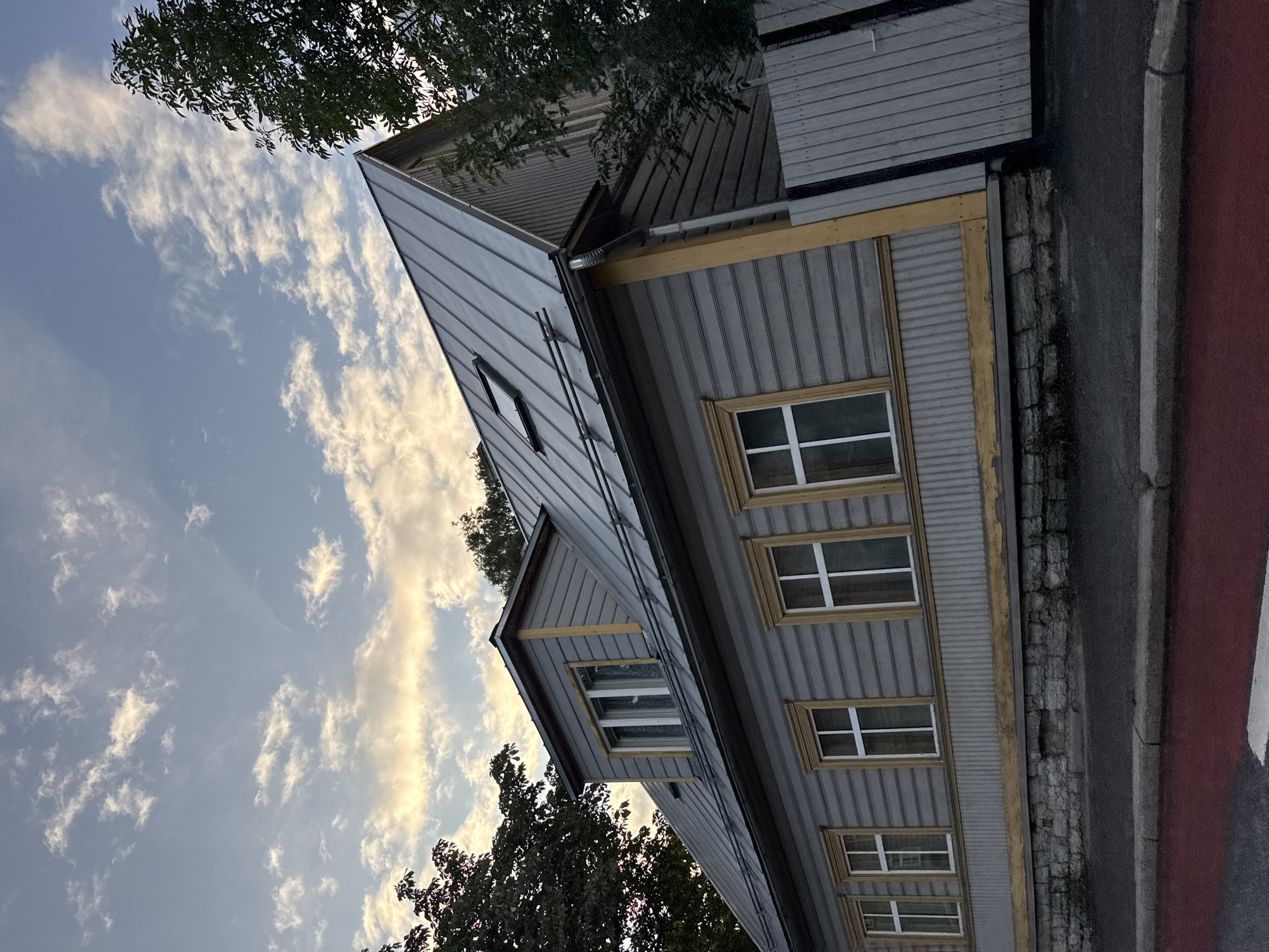 The image shows a two-story wooden house with a metal roof on a street in Tallinn, Estonia. The house is grey with yellow trim around the windows and doors. There are several windows on the front of the house, and a dormer window on the roof. A wooden fence is visible on the right side of the house. The sky is partly cloudy, with the sun shining through the clouds. The street in front of the house is paved with asphalt and has a red lane marking. The house appears to be old, with some signs of wear and tear. The overall impression is of a well-maintained, but aged, residential building in an urban setting.