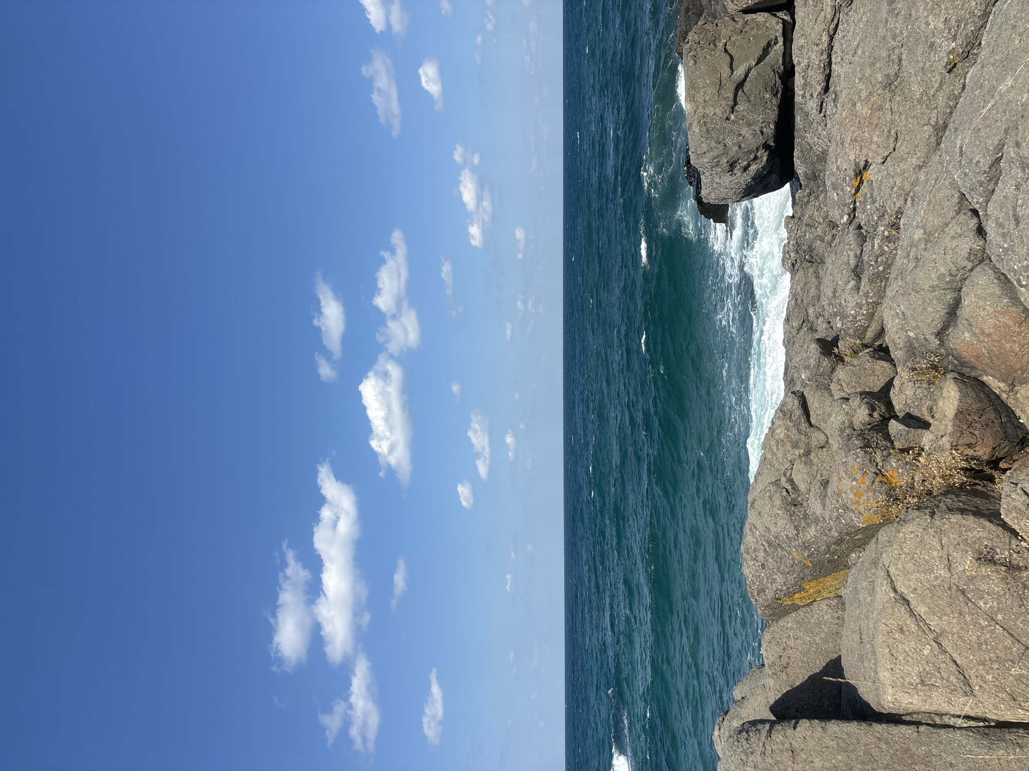 The image depicts a rocky coastline under a bright blue sky with scattered white clouds. The sea is a deep turquoise with white-capped waves crashing against the shore. The foreground is dominated by large, grey rocks, some of which are covered in patches of orange lichen. The environment appears to be a natural, undeveloped coastal area. The scene is likely located in the vicinity of Sinemorets, Bulgaria, known for its rugged coastline and clear waters. There are no people or man-made objects visible in the image.