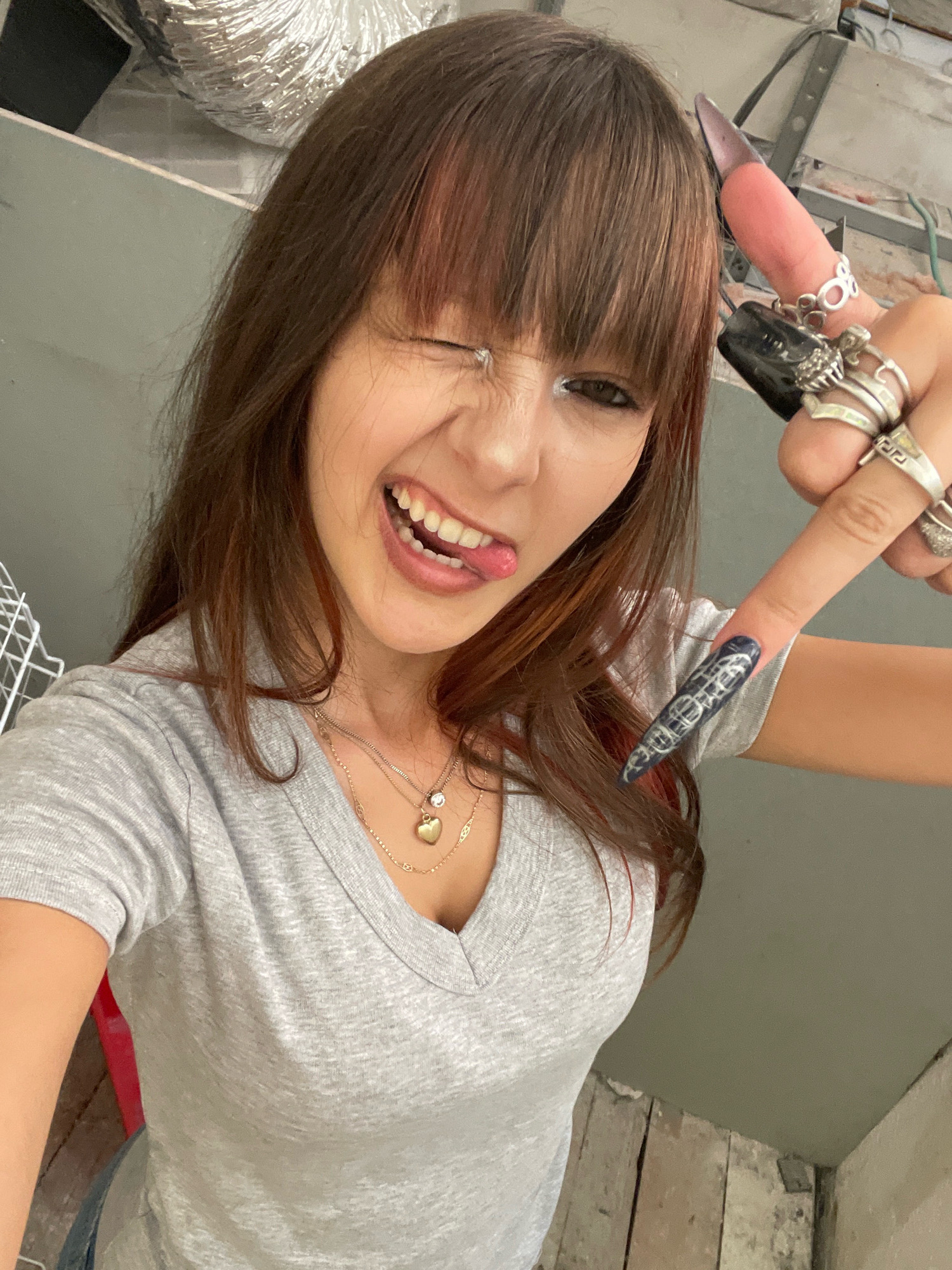 A young woman with long brown hair and bangs is taking a selfie. She is wearing a grey V-neck t-shirt and has on multiple rings on her fingers, some of which have elaborate designs. Her fingernails are long and pointed, with one appearing to have a metallic sheen and the other a dark color with a design. She is winking and sticking her tongue out, with a playful expression. The background appears to be an indoor space with exposed pipes and ductwork, suggesting an unfinished or industrial setting. The image is cropped tightly around the woman, focusing on her face and upper body. There are no discernible landmarks or elements that would indicate a specific location within Raanana, Israel.