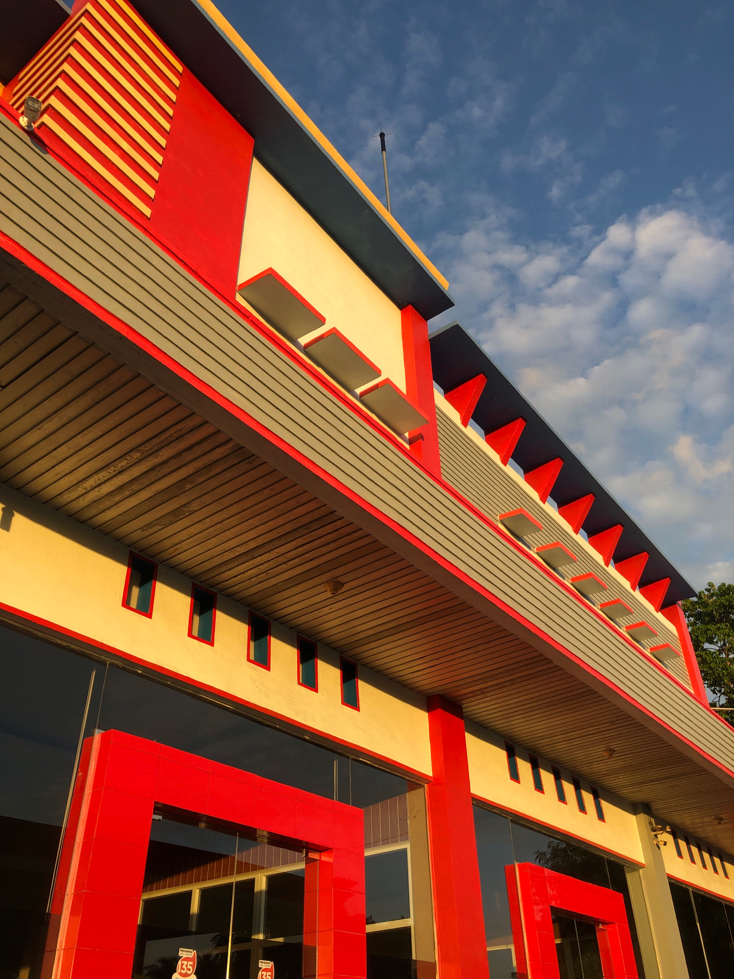 The image shows a low-angle exterior shot of a modern building with a striking red, gray, and yellow color scheme against a blue sky with scattered clouds. The building features geometric architectural elements, including a red entranceway with a cubist design, a series of small, rectangular windows along the upper facade, and a distinctive roofline with red triangular accents and recessed gray panels. The upper portion of the building has a section with diagonal yellow and red stripes. The time of day appears to be late afternoon or early evening, as evidenced by the warm, golden light illuminating the building's surfaces. Reflections of the building and the sky are visible in the large glass windows on the ground floor. The building is likely a commercial establishment, possibly a gas station or a retail outlet, given the "PERTAMINA" logo visible on signage reflected in the windows. This building is located in Lhokseumawe, Indonesia. There are no people visible in the image.