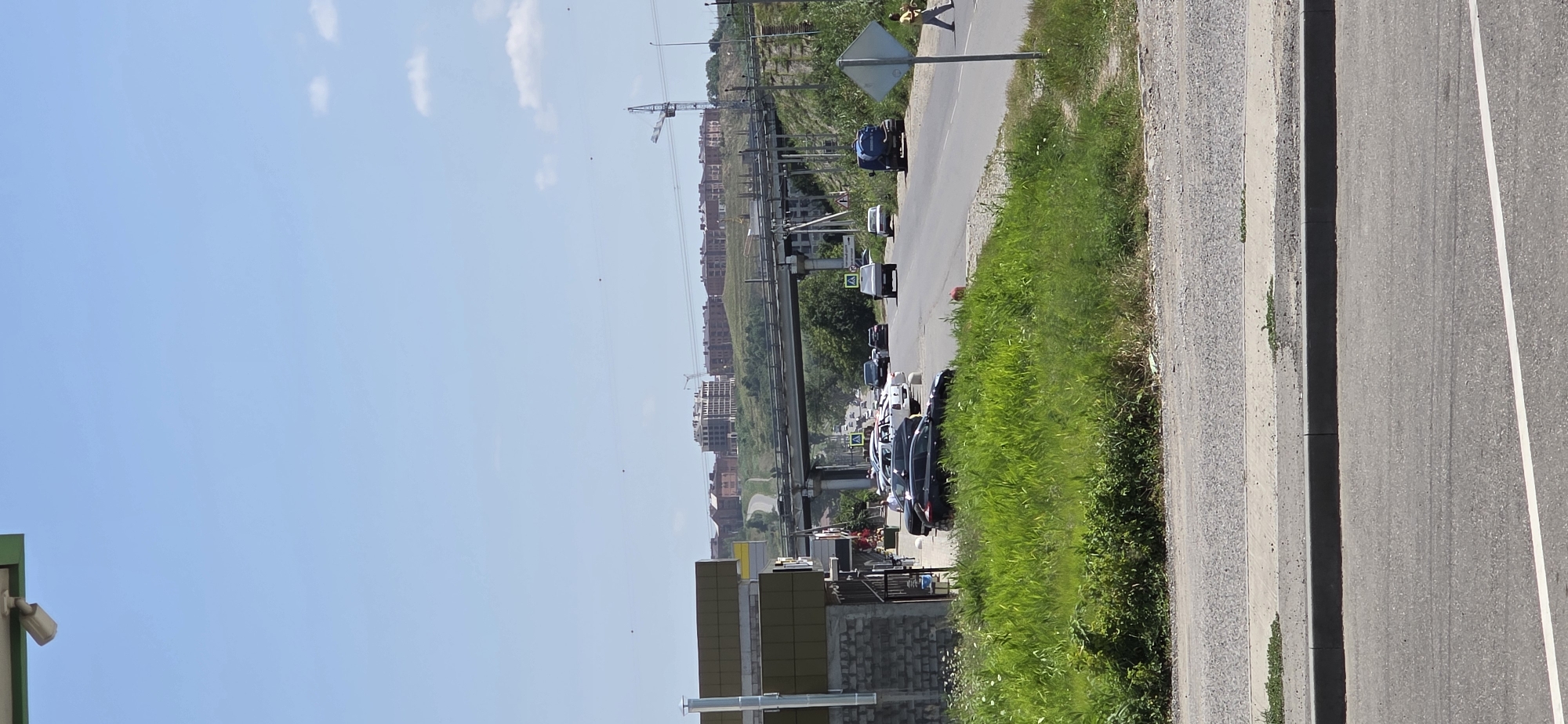 The image depicts a bright, sunny day in Inozemtsevo, Russia. In the foreground, a two-lane asphalt road with a white line marking the center is visible. To the left of the road, a patch of vibrant green grass and weeds grows next to a gravel shoulder and a concrete curb. Further left, a modern building with a dark green roof and gray brickwork stands adjacent to the road. Beyond this building, a few cars are parked.
The middle ground features a busy road with several vehicles in motion, including a black SUV, a white sedan, and a blue truck with a water tank. The road curves to the right, leading under an elevated structure, likely a bridge or overpass, which spans across the scene. The area surrounding the road is characterized by a mix of green vegetation, including tall grass and bushes, and dirt or gravel patches.
In the background, a city skyline with numerous multi-story buildings, some appearing to be under construction with cranes visible, is situated on a gently sloping hill. The buildings are primarily brick-colored with modern architectural designs. The sky above is a clear, pale blue, with a few wispy white clouds scattered across it.
A lone pedestrian, dressed in a yellow shirt and dark pants, is seen walking across the road from right to left, near a diamond-shaped blue and white road sign. Another person is partially visible on the far right side of the image, also walking. The overall scene suggests a suburban or semi-urban environment with ongoing development.