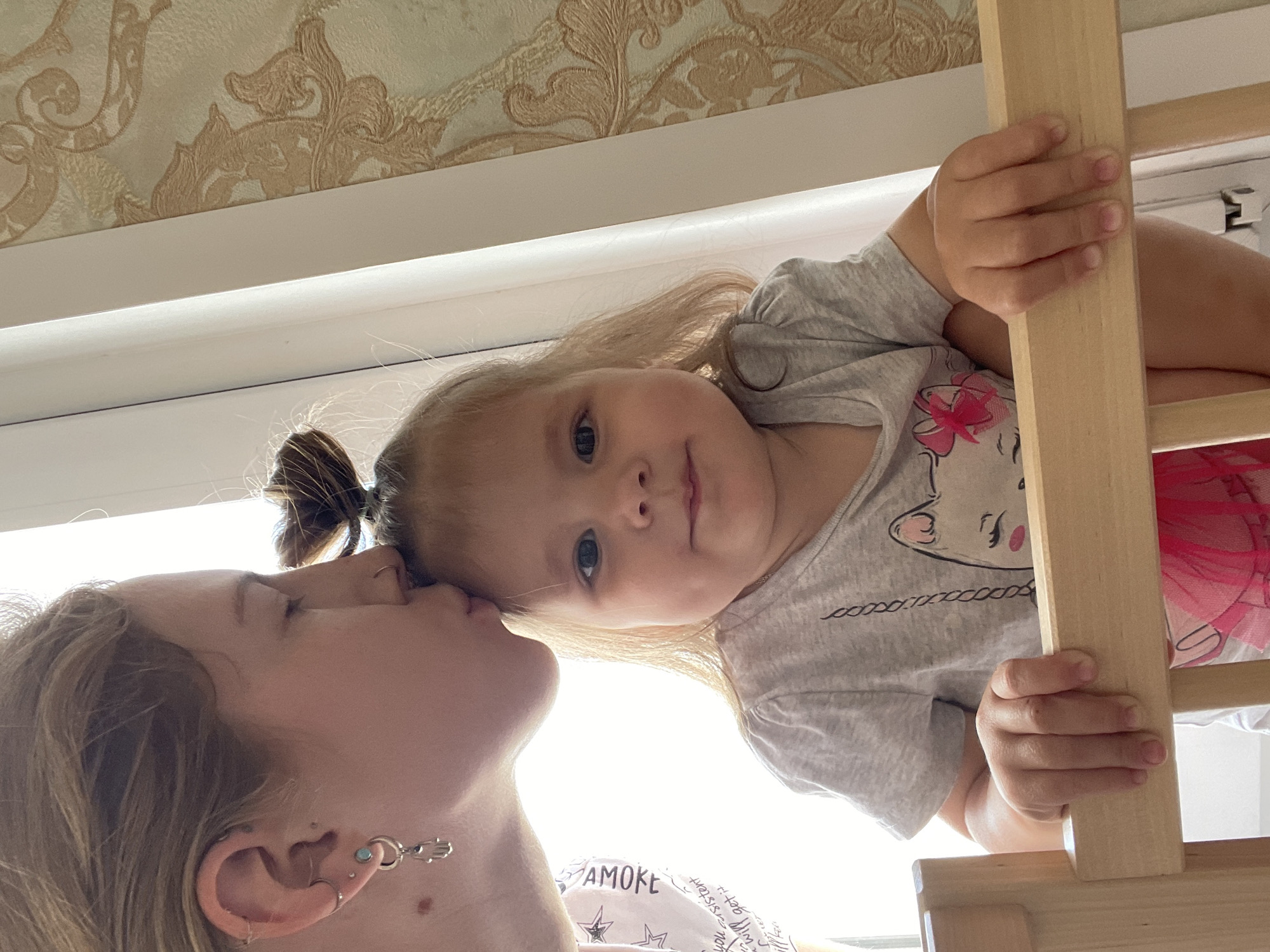 The image captures a tender, close-up moment between two individuals in an indoor setting in Izhevsk, Russia. The photo is oriented with the subjects lying on their sides.
On the left, an adult or older child with light brown hair, partially styled into a bun or ponytail, is seen in profile. Their eyes are closed, and they are leaning in to kiss or nuzzle the forehead of the younger child. They wear multiple ear piercings, notably a silver Hamsa hand charm earring and several silver hoops/studs. Part of their light-colored top is visible, with the faint word "AMOKE" printed on it, and further down, the words "you will get" and "sister consistent".
To the right and slightly above, a young child, likely a toddler or preschooler, with blonde hair, looks directly at the camera with wide blue or grey eyes and a calm expression. Their hair is parted and frames their face, with a small ponytail visible at the top of their head. They are wearing a light grey t-shirt featuring a cartoon graphic of a white cat-like character with pink cheeks and a pink bow/flower. Faint, repeated text is visible below the cartoon. The child's right hand grips a vertical wooden beam, part of a light-colored wooden structure, possibly a bunk bed or play ladder, which is visible on the right side of the frame. Behind the child, a section of pink, translucent fabric, possibly from a tutu, is visible.
The background features a bright, overexposed light source, likely a window, creating a halo effect around the subjects. Above this, a horizontal white beam runs across the frame. At the very top, an ornate patterned wallpaper with swirling beige and gold floral or rococo designs on a lighter background is visible, suggesting a cozy home interior. The strong light indicates daytime. The scene conveys warmth and affection.