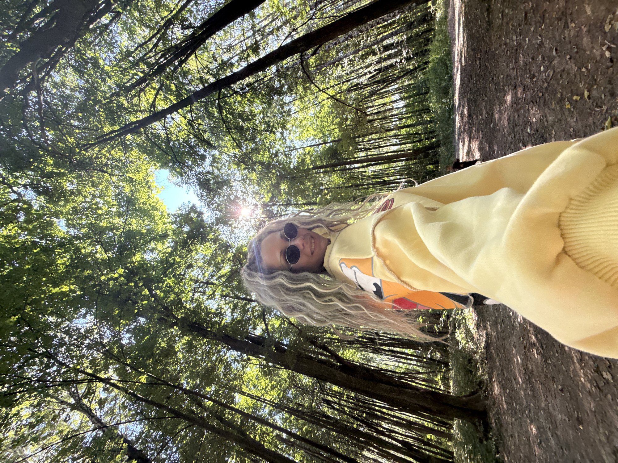 A low-angle, medium shot captures a woman with long, wavy blonde hair, wearing sunglasses and a yellow hoodie, posing for a selfie in a sun-dappled forest. The trees, with their tall, slender trunks, create a dense canopy overhead, allowing sunlight to filter through the vibrant green leaves. The forest floor is covered in fallen leaves and dappled shadows. The woman's face is turned towards the camera, with a slight smile. The image appears to be taken on a clear day, with the sun shining brightly. The location is identified as a forest in Rusanovo, Russia.