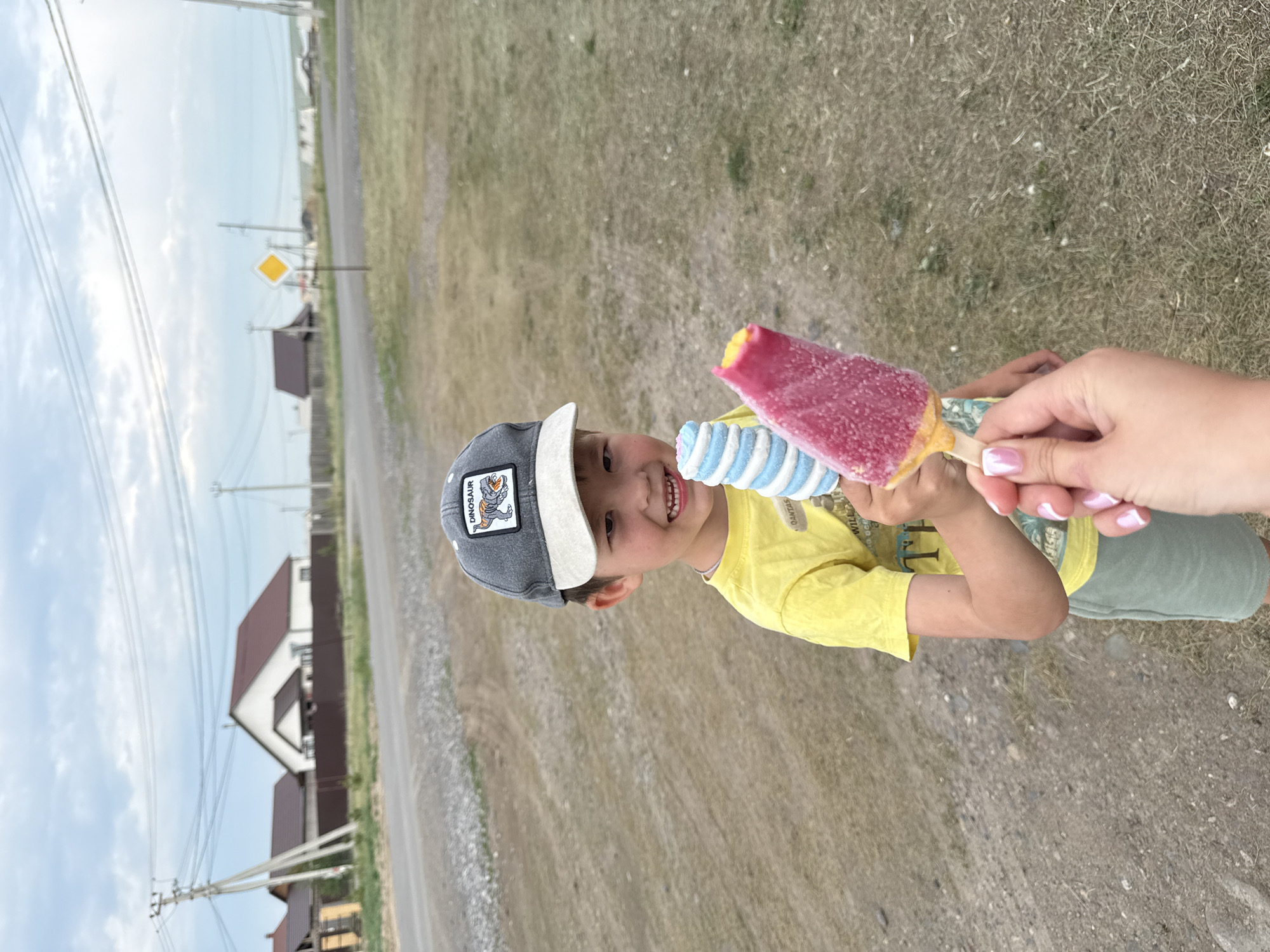 In Bely Yar, Russia, a young boy wearing a gray baseball cap with a dinosaur patch and a yellow t-shirt stands outdoors, holding two popsicles. One is a bright pink rectangular popsicle with a yellow bottom, and the other is a blue and white swirled cylindrical popsicle. The boy is smiling and looking at the camera. A woman's hand with manicured nails holding a third popsicle is visible in the foreground, reaching towards the boy. The background shows a dirt road, dry grass, and houses with pitched roofs, characteristic of a rural or suburban area. Utility poles and wires are also visible in the distance. The sky is overcast.
