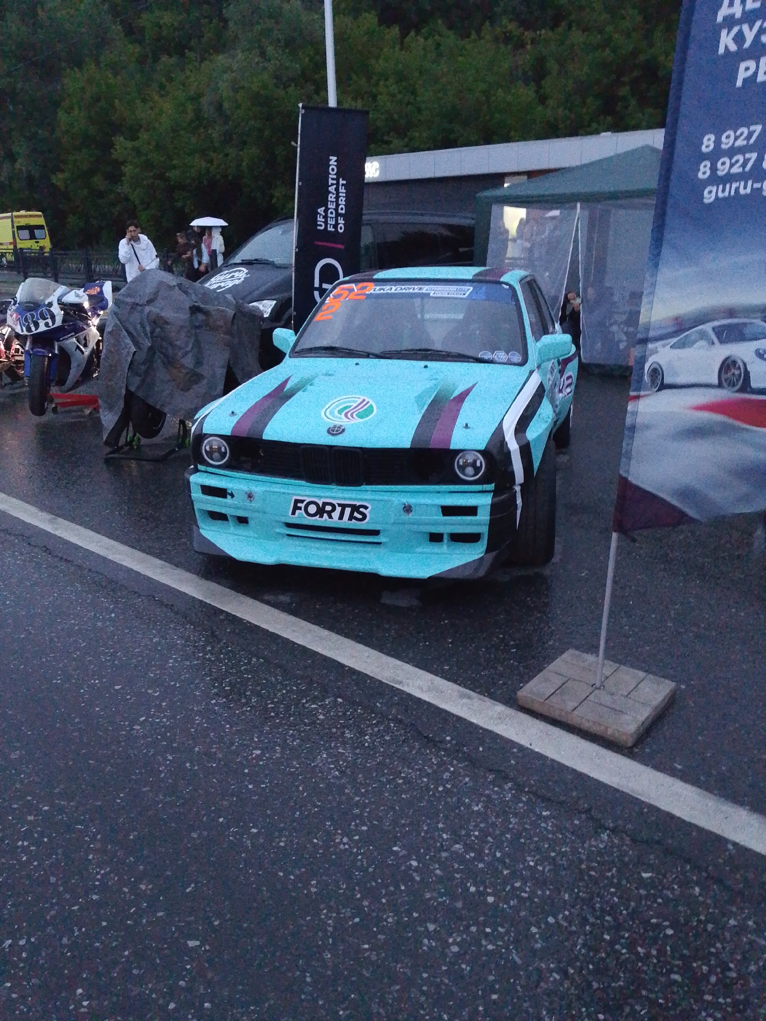 The image captures a vibrant motorsport event, possibly a drift competition, in Bulgakovo, Russia. The focal point is a heavily modified, light blue BMW E30 race car, adorned with racing decals and sponsor logos, including "FORTIS" prominently displayed on its front bumper. The car is positioned on a wet asphalt track, indicated by the reflection and speckles on the surface.
To the left of the car, a blue and white racing motorcycle with the number "189" is visible. In the background, a dark-colored SUV, a yellow utility vehicle, and a group of spectators under an umbrella suggest a gathering of enthusiasts. A large black banner with white text, partially obscured by the race car, features the words "UFA FEDERATION OF DRIFT," confirming the event's nature and location.
Further to the right, a large banner displaying an image of a white Porsche and Russian text, including phone numbers and the word "GURU," is unfurled. The environment appears to be an outdoor venue, possibly a track or event grounds, with trees forming a dense backdrop. The weather seems overcast or rainy, contributing to the wet conditions. The presence of people, vehicles, and event-specific signage indicates an active and engaging motorsport occasion.