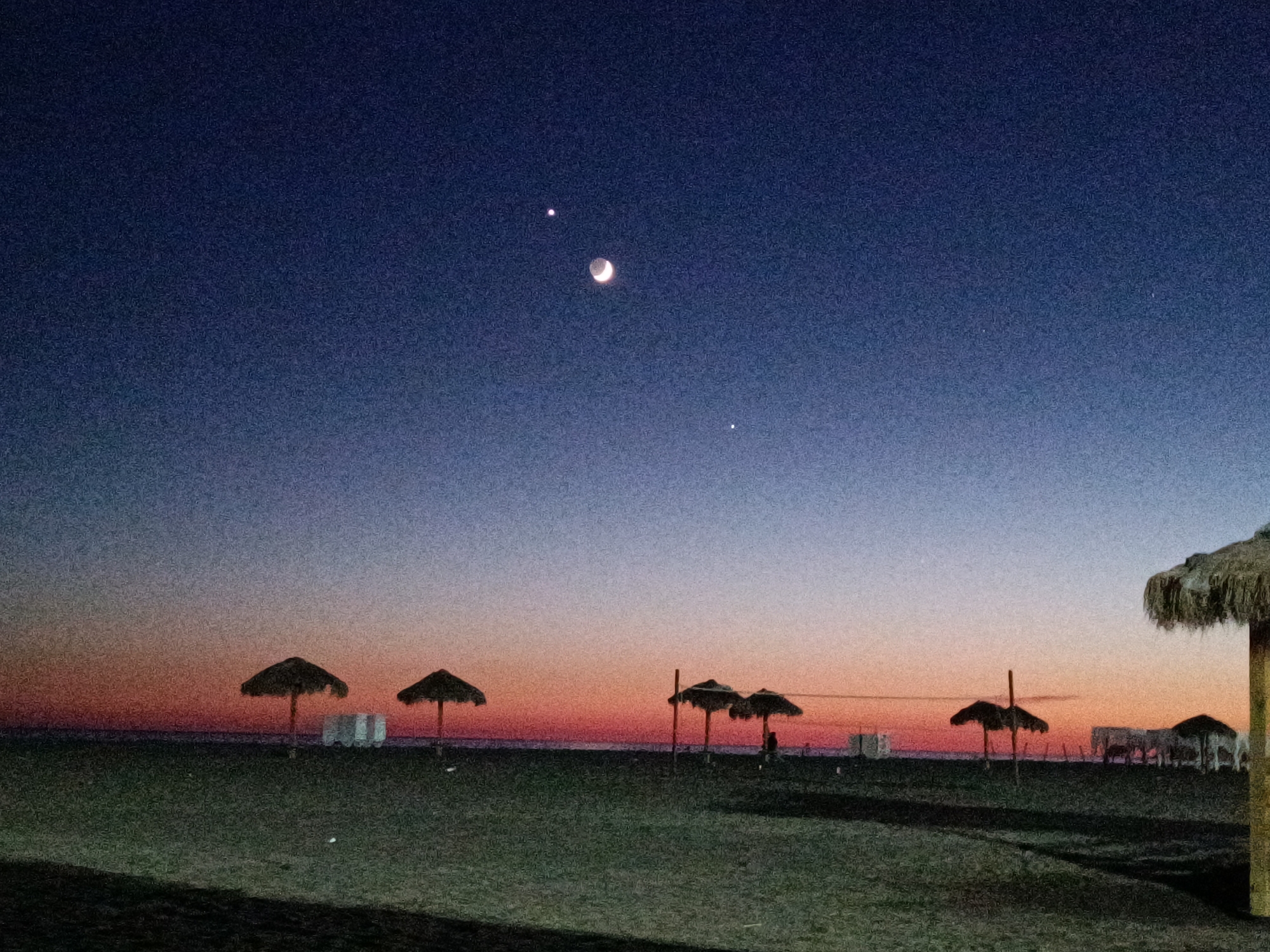 The image depicts a beach scene at twilight. The sky transitions from a deep blue at the top to shades of purple, pink, and orange near the horizon. In the sky, a crescent moon is visible along with two bright celestial bodies, likely planets. The foreground consists of a sandy beach with scattered umbrellas, some with thatched roofs, and white structures that might be beach chairs or cabanas. A single, tall umbrella with a thatched roof is prominent on the right side of the frame. There are no people clearly visible in the image, though shadows suggest some presence. The overall atmosphere is calm and serene, characteristic of a beach at the end of the day.