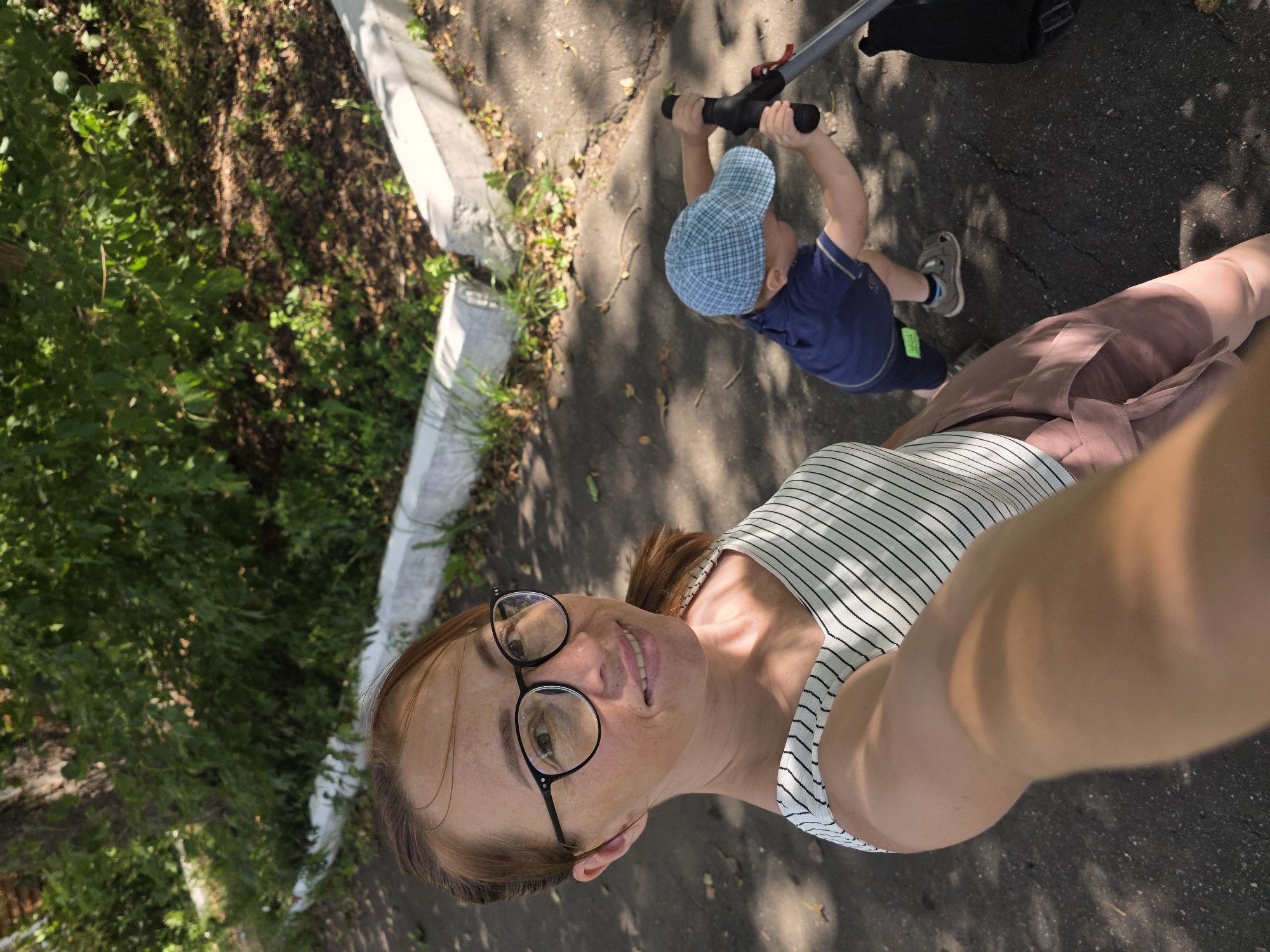 In an outdoor setting, a woman with reddish-brown hair pulled back and wearing glasses and a striped tank top smiles at the camera, holding it to take a selfie. She is wearing a dusty rose colored skirt. To her right, a young child wearing a blue plaid cap and a blue shirt is pushing a stroller. The scene appears to be a park or wooded area with a paved path and some concrete curbing visible. The presence of trees and greenery suggests a natural environment within a populated area. The lighting indicates it is daytime, with dappled sunlight creating shadows on the ground. The image has a candid feel, capturing a moment of interaction between a parent and child outdoors, possibly in the city of Chaikovsky, Russia, given the provided location.