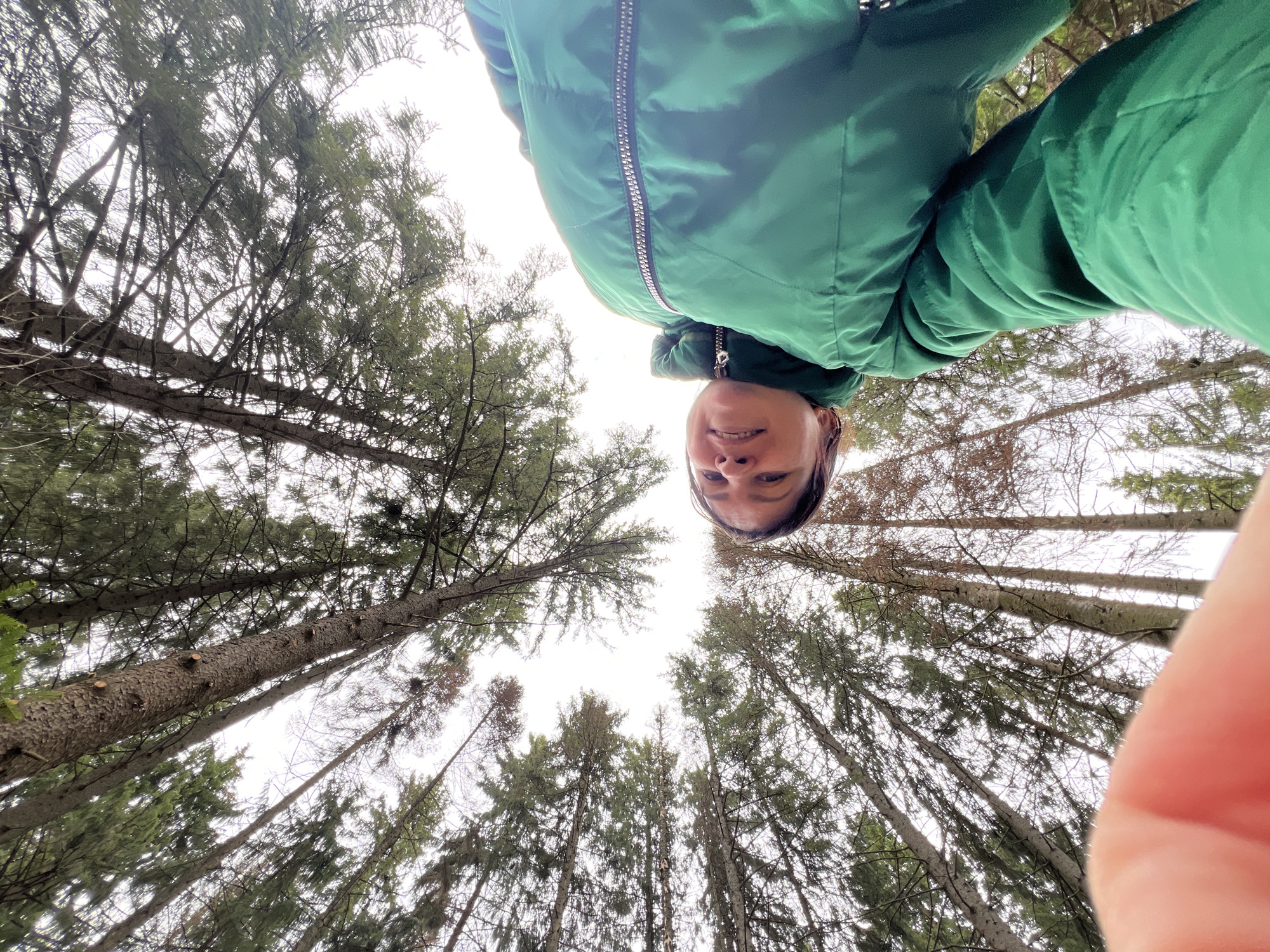 A low-angle shot captures a person wearing a green zip-up jacket, standing amongst tall trees in what appears to be a forest. The location is potentially in Protasy, Russia, based on the metadata. The individual is looking down at the camera. The background consists of numerous trees reaching up towards the sky, with a slightly overcast sky visible between the branches. A finger is visible in the frame's bottom right corner.