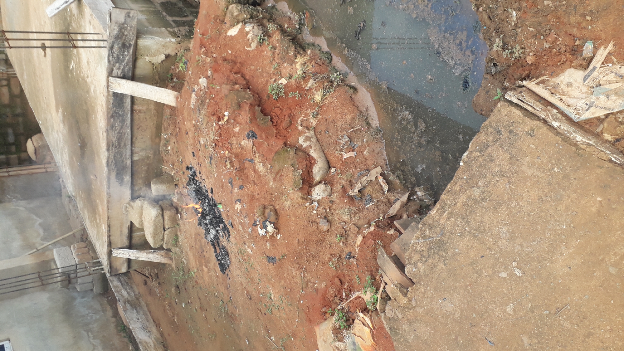 The image depicts an outdoor scene in Nkpor, Nigeria, characterized by a construction site with elements of neglect and disarray. A small fire burns on a pile of reddish-brown soil, with ash scattered around it. Adjacent to the fire is a pool of murky, stagnant water. The ground is uneven and appears to be a mixture of soil, debris, and some construction materials.
In the background, a partially constructed building is visible, with exposed rebar and concrete slabs. There are also stacks of rough-hewn stones and some wooden planks leaning against structures. The overall impression is one of an unfinished and untended construction area, with signs of recent activity (the fire) and ongoing deterioration. The environment appears to be a typical urban or semi-urban setting within Nkpor, likely an area undergoing development but not currently under intensive, organized construction. No individuals are present in the image.
