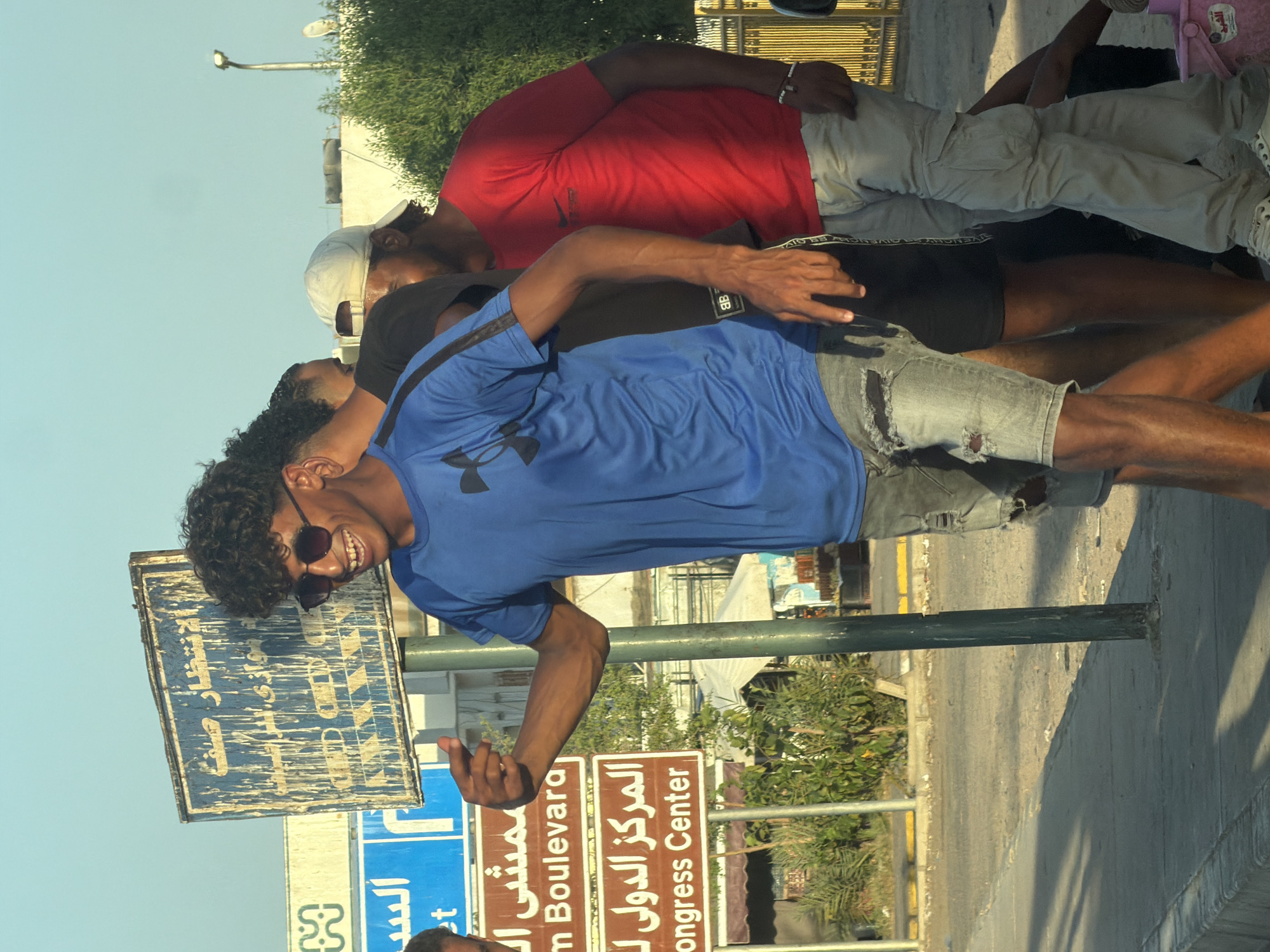 The image shows a group of young men standing on a sidewalk in Sharm El Sheikh, Egypt. The man in the foreground, wearing a blue t-shirt and ripped denim shorts, is smiling and gesturing with his left hand. Behind him, other men are visible, one in a red t-shirt and light-colored pants, and another in a black shirt. Several signs are present in the background, including one with Arabic text that appears to be a street or building name, and another in English that reads "Congress Center" and "Am Boulevard." The environment appears to be urban, with a street, sidewalk, and some greenery visible. The lighting suggests it is daytime, with the sun casting shadows on the ground.