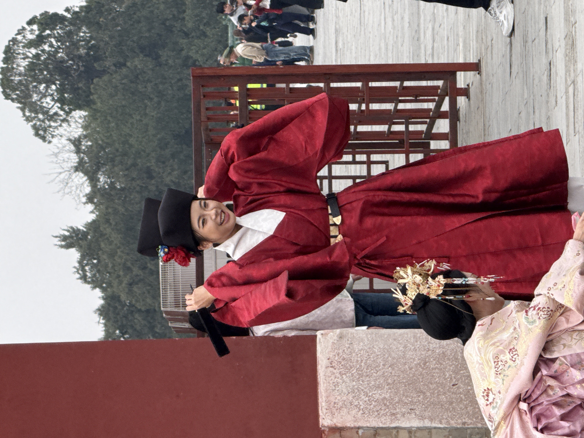 The image presents an outdoor scene within Dongcheng District, China. A young East Asian female stands centrally, smiling towards the viewer, dressed in a red traditional Chinese robe with wide sleeves over a white inner garment. She wears a black flat-topped hat adorned with red flowers and blue elements, holding the sides of the hat with both hands. A black belt with gold-colored decorative plates is cinched at her waist. In the lower left foreground, another East Asian female is depicted kneeling or bending low, wearing a light pink traditional robe with gold and floral patterns. Her dark hair is styled with an elaborate golden headdress featuring multiple dangling ornaments.
To the immediate left of the central figure is a prominent red architectural wall with a light-colored stone base. On the right, a dark reddish-brown metal gate with vertical bars acts as a barrier. Beyond this gate, several individuals in contemporary casual clothing, including adults and a child, are visible in the mid-ground, appearing to observe the scene. One male figure wears a beige jacket and blue jeans, while another has a green cap. The ground consists of light gray paving stones or tiles. The far background is dominated by a dense canopy of dark green trees under an overcast sky.