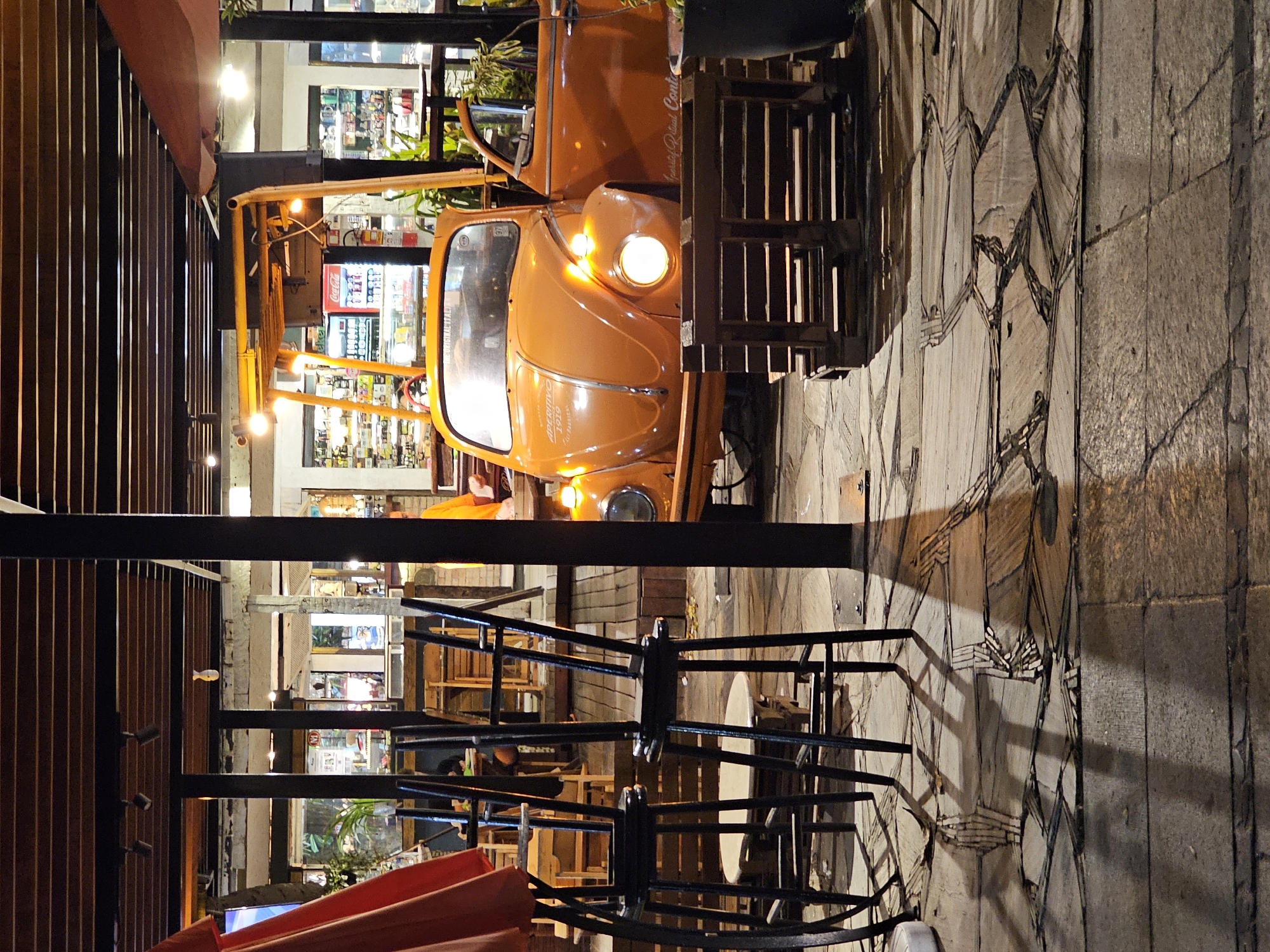 The scene depicts a nighttime view of an outdoor patio area in Armação Dos Búzios, Brazil, fronting a commercial establishment. A prominent orange vintage Volkswagen Beetle occupies the mid-ground, oriented slightly left of center with its right-side door open and its front right headlight illuminated. The hood displays "APERITIVO 1919" and "THE ORIGINAL ITALIAN" text, while "Quality Ritual Conto" is visible on the open door. A yellow metal framework extends above the car's rear section. In the left foreground, several dark-colored bar stools are inverted and stacked on a round, light-colored table, partially obscured by a red umbrella or awning structure. A stack of wooden pallets rests on the ground to the right of the car's front bumper, adjacent to a dark potted plant. The ground is paved with irregularly shaped stone tiles featuring a mosaic-like pattern, transitioning to wooden decking further back. Multiple dark vertical pillars support a slatted wooden overhead canopy containing recessed lighting. The background reveals the interior of the establishment through glass windows, showcasing illuminated shelves stocked with bottles and merchandise, including a red Coca-Cola refrigerator. No clearly discernible individuals are present within the frame.