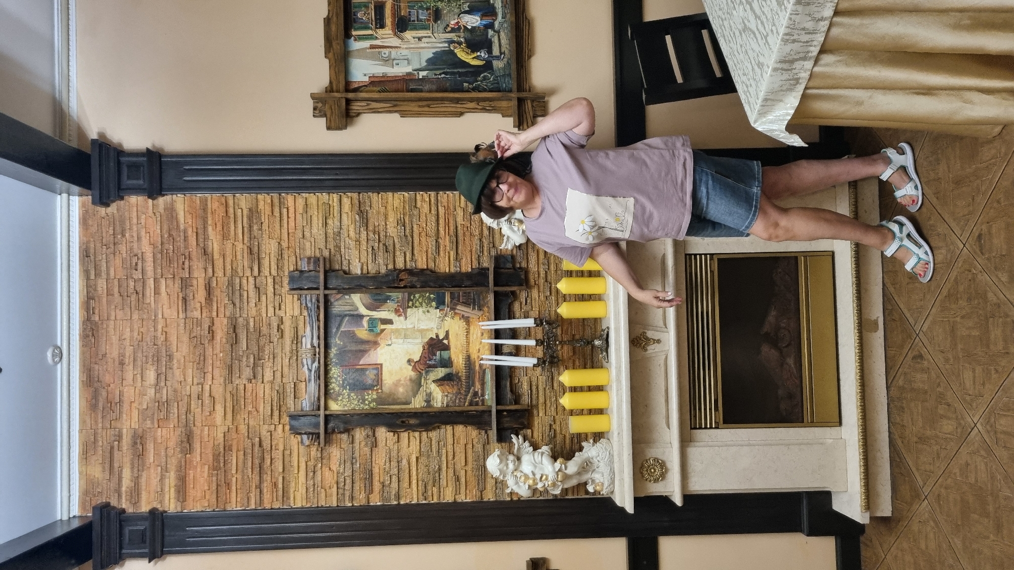 A woman wearing a green hat, a purple t-shirt with a daisy print, and denim shorts stands next to a fireplace. She is touching the brim of her hat. The fireplace is made of marble and has an electric insert with a gold trim. On the mantlepiece are a candelabra with four white candles, seven yellow pillar candles, and two white cherub statues. Above the mantlepiece, there is a painting in a wooden frame depicting a person sitting in a room. The wall behind the fireplace is made of stacked stone. To the right of the fireplace, there is a table covered with a beige tablecloth. In the background, a framed painting of a European street scene is visible on the wall. The scene appears to be indoors, possibly in a restaurant or a home, given the presence of a fireplace and dining table. The style suggests a location in Chernivtsi, Ukraine, due to common decorative elements found in establishments there.