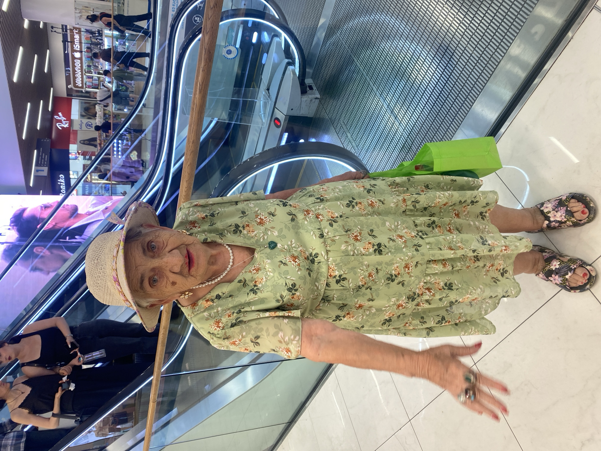 An elderly woman wearing a light-colored straw hat and a light green floral dress stands on a white tiled floor next to a moving escalator. She has a pearl necklace and a green pendant around her neck. Her right hand is extended, showing rings on two fingers. She is carrying a bright green shopping bag in her left hand. The escalator has blue LED lights along its sides. In the background, there are various shop signs, including "Roniko" and "Ray-Ban," suggesting the location is a shopping mall. Other shoppers are visible on the escalator and in the background shops. The scene appears to be in Tbilisi, Georgia, as indicated by Georgian text on some of the shop signs.