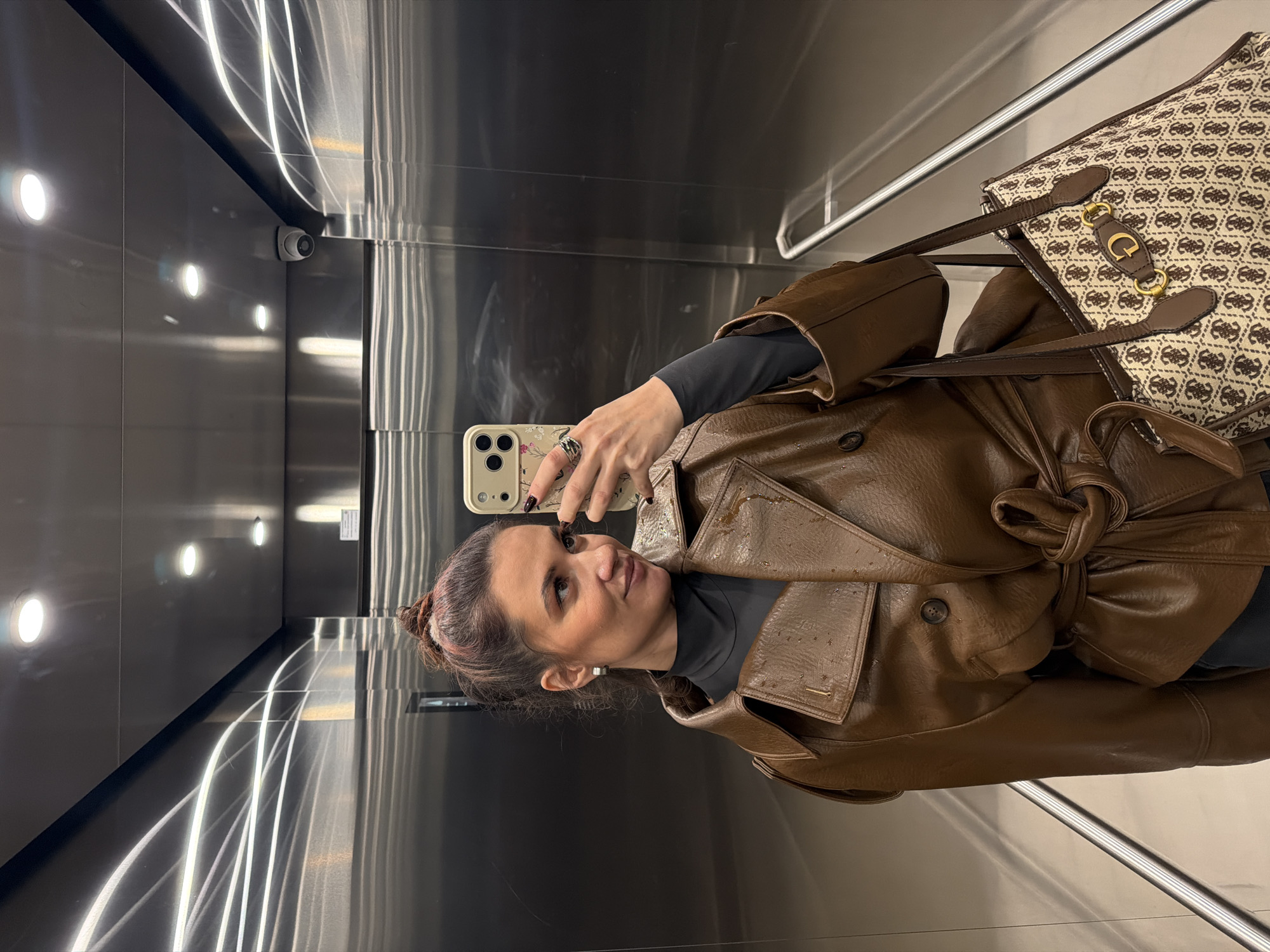 A female individual is depicted in a mirror selfie taken inside a modern elevator. She stands angled slightly to her right, with her head turned to her left, looking into the smartphone held in her right hand, which is capturing her reflection. Her dark hair, with subtle reddish highlights, is pulled back into a high bun. She is dressed in a dark, long-sleeved top and a brown, textured trench coat-style jacket, cinched at the waist with a self-tie belt. The jacket features a wide collar and a visible button closure. She wears silver hoop earrings, dark nail polish, and a ring on her right hand. A large tote bag with a beige and brown repeating monogram pattern and a prominent gold "G" logo with chain accents hangs from her left shoulder.
The elevator interior features highly reflective brushed metallic walls and ceiling, likely stainless steel, which display abstract light reflections and the subject's mirrored image. Three circular recessed lights are visible on the ceiling, along with a white dome-style security camera. A metallic handrail extends along the lower portion of the left wall. The setting is an interior elevator within a modern commercial or public building in Belgrade, Serbia.