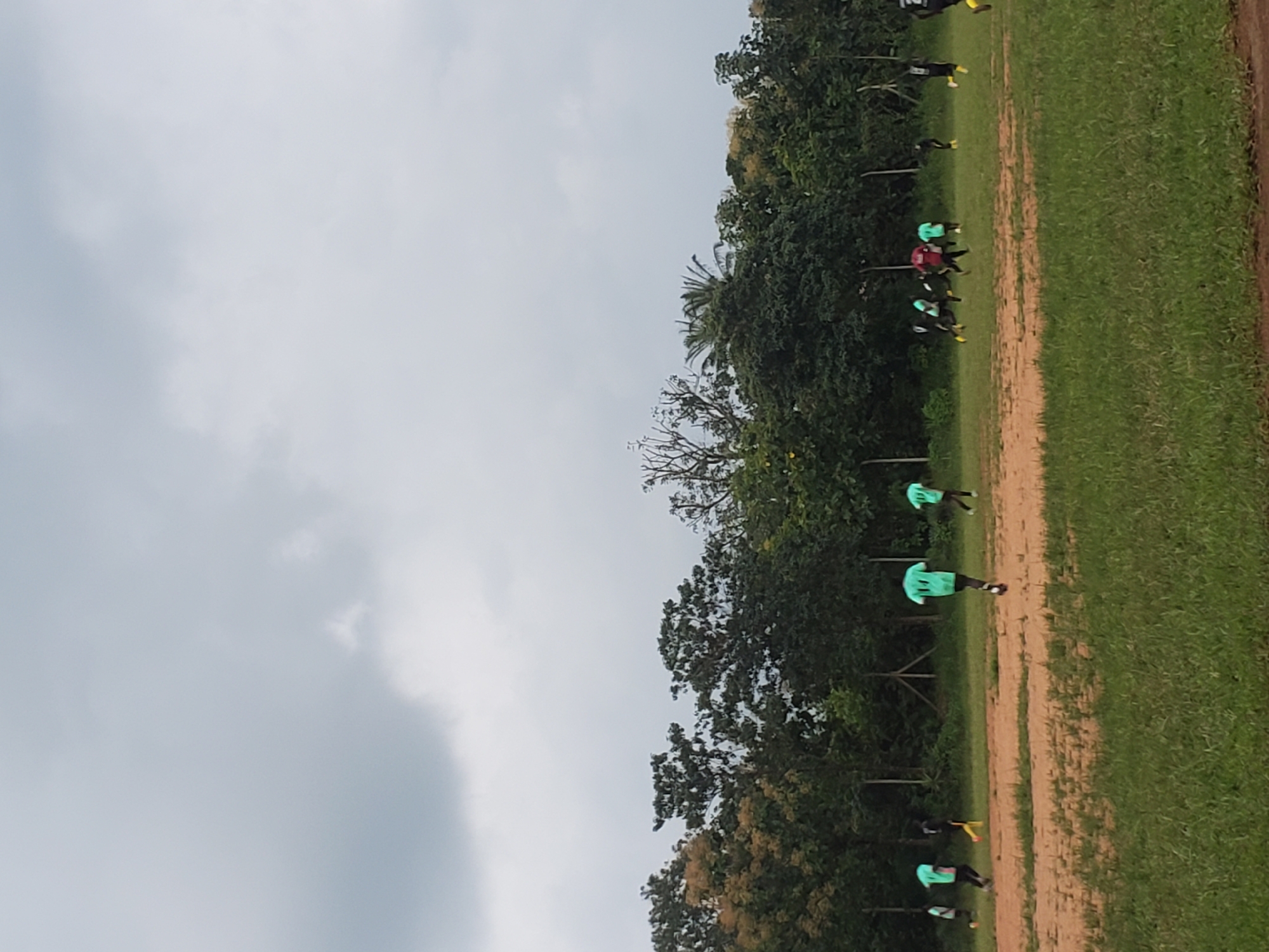 The image depicts a football practice session taking place on a dusty field surrounded by lush green trees and vegetation, under a cloudy sky. The field appears to be a local sports ground in Miango, Nigeria. Several individuals, likely young men, are engaged in playing football. Most of them are wearing bright green t-shirts, while a few are in dark attire. Some players are actively running and kicking the ball, indicating a game in progress. The overall setting suggests a casual sporting event in a natural, outdoor environment.