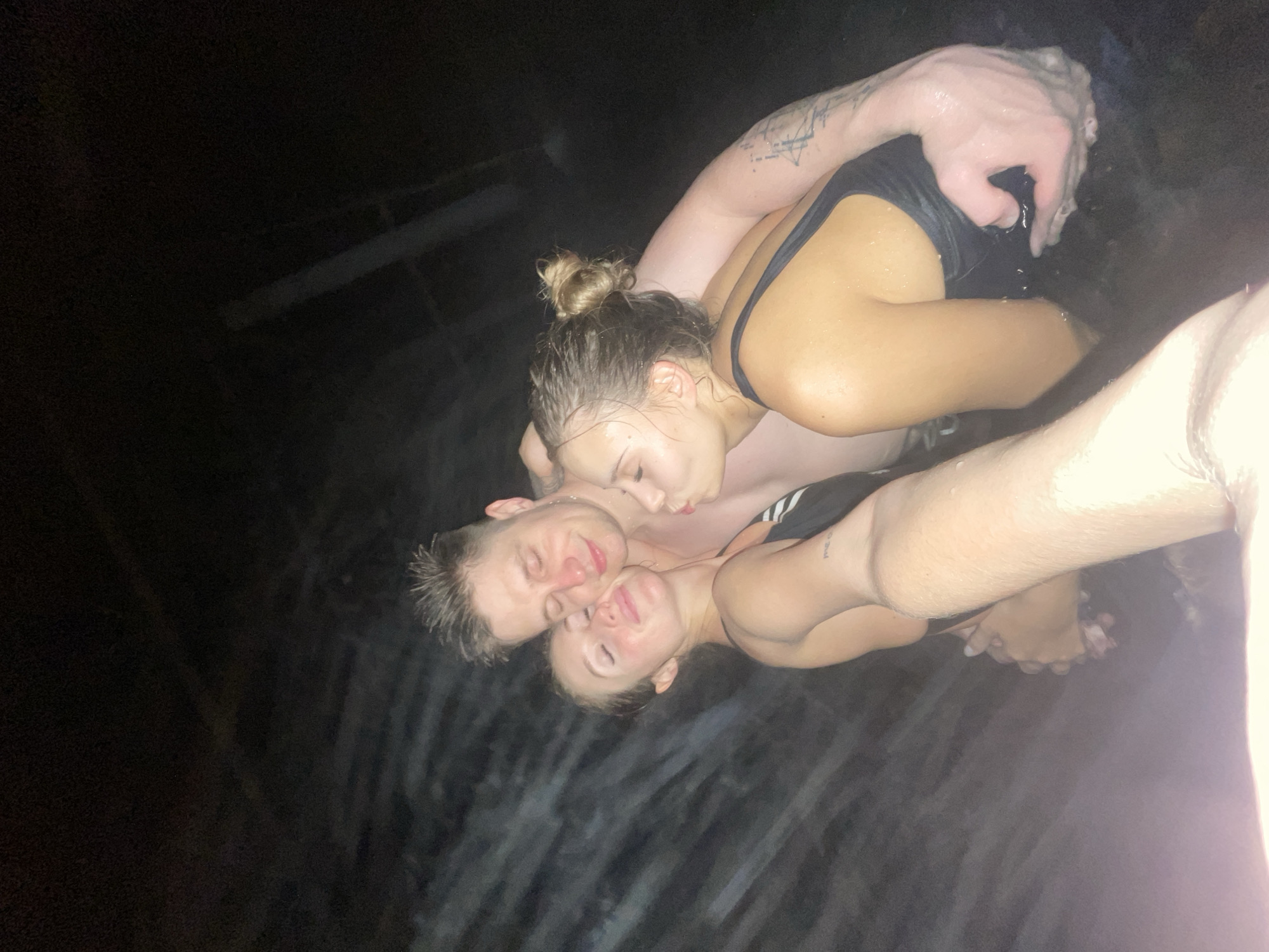 Three individuals are captured in what appears to be a body of water, potentially a hot tub or pool, situated in an environment that seems to be dimly lit, almost as if it were night. The leftmost individual, a male, is seen with his eyes closed, his head tilted slightly, and his face in close proximity to another person, potentially suggestive of an intimate embrace. Slightly above him, a female is also with closed eyes, who appears to be kissing his neck. The third individual is barely visible but can be seen holding the other two individuals. All three are wearing swimwear. The lack of light and the submerged environment add to the mystery of the scene. The location is Unknown, Unknown.