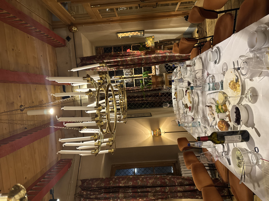 The image depicts an empty dining room in Tbilisi, Georgia, following a meal. A long, white-tableclothed dining table occupies the foreground, surrounded by numerous unoccupied orange-brown upholstered chairs. The table is covered with the remnants of a meal, including partially consumed plates of food such as roasted items, bread, a dark berry-like dish, green salad/dip, and desserts. An empty red wine bottle, multiple used wine glasses with red wine residue, clear water bottles (one identified as Borjomi), teacups, saucers, and teapots are scattered across the table.
Overhead, a large, multi-tiered brass chandelier featuring numerous unlit white taper candles hangs from a wooden ceiling with prominent red-painted beams. Additional illumination comes from a single ceiling bulb and two floor lamps with patterned shades, casting warm light throughout the room. The walls are light-colored. Multiple windows with leaded glass panes are visible, draped with heavy, patterned red and brown curtains. Outside the windows, it is dark. A potted plant sits on a stand near one window, and a wooden door with glass panels is situated on the right side of the room. No individuals are present in the scene.
