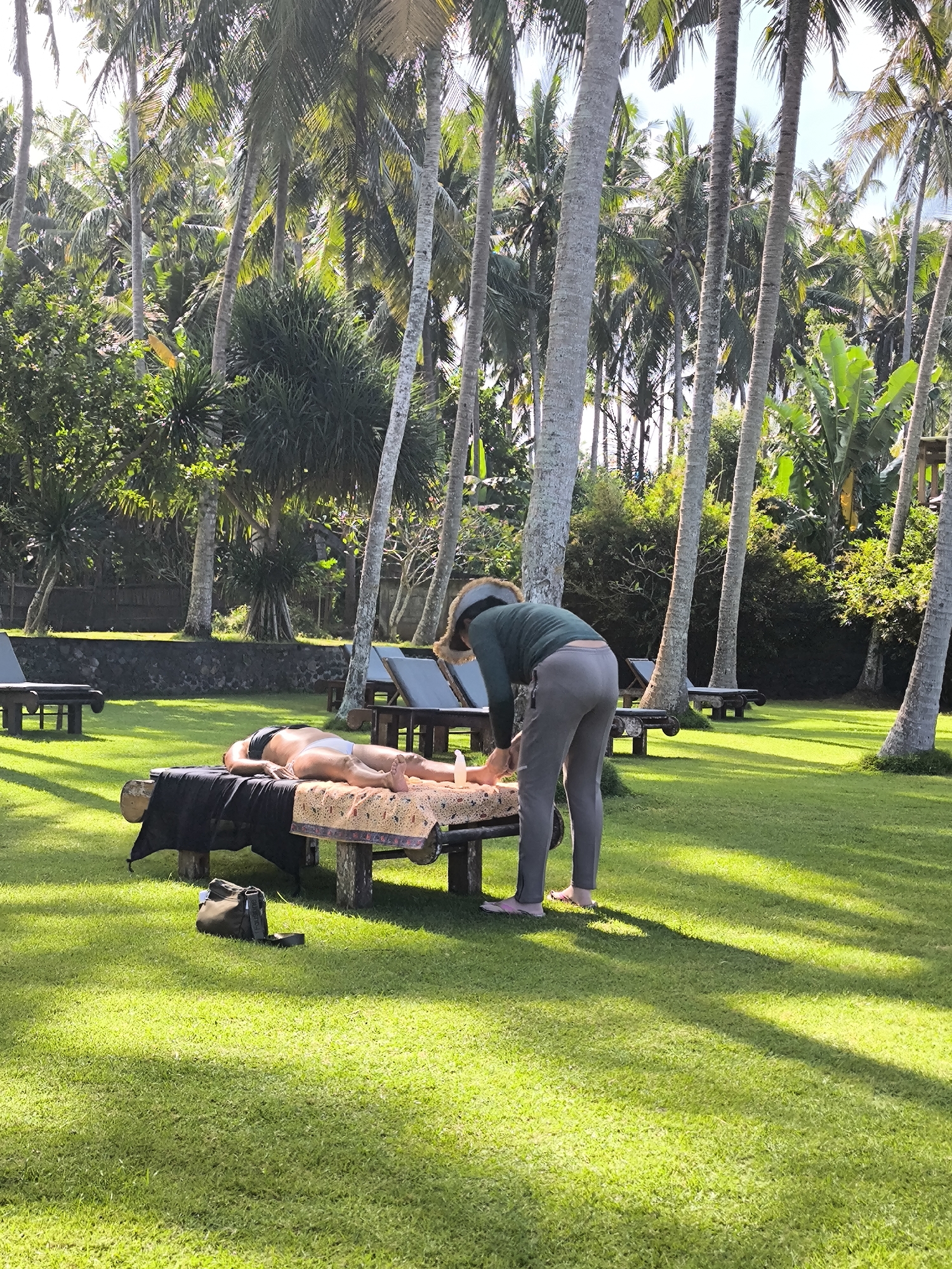 The image depicts an outdoor massage scene in a lush, tropical garden environment. The location is in Karangasem, Indonesia, characterized by the abundant presence of tall coconut palm trees and dense green foliage. In the foreground, a person is lying face down on a massage table, which is supported by rustic wooden logs. The person is wearing white underwear and appears to be receiving a massage. A masseuse, wearing a green long-sleeved shirt and grey pants, is attending to the person, with their hands positioned near the person's back. A patterned cloth is draped over the massage table, and a black cloth hangs from the side. Next to the massage table, on the vibrant green grass, a dark-colored bag is placed. In the background, other massage tables and lounge chairs are visible, suggesting a spa or resort setting. Sunlight filters through the palm trees, casting dappled shadows on the grass.