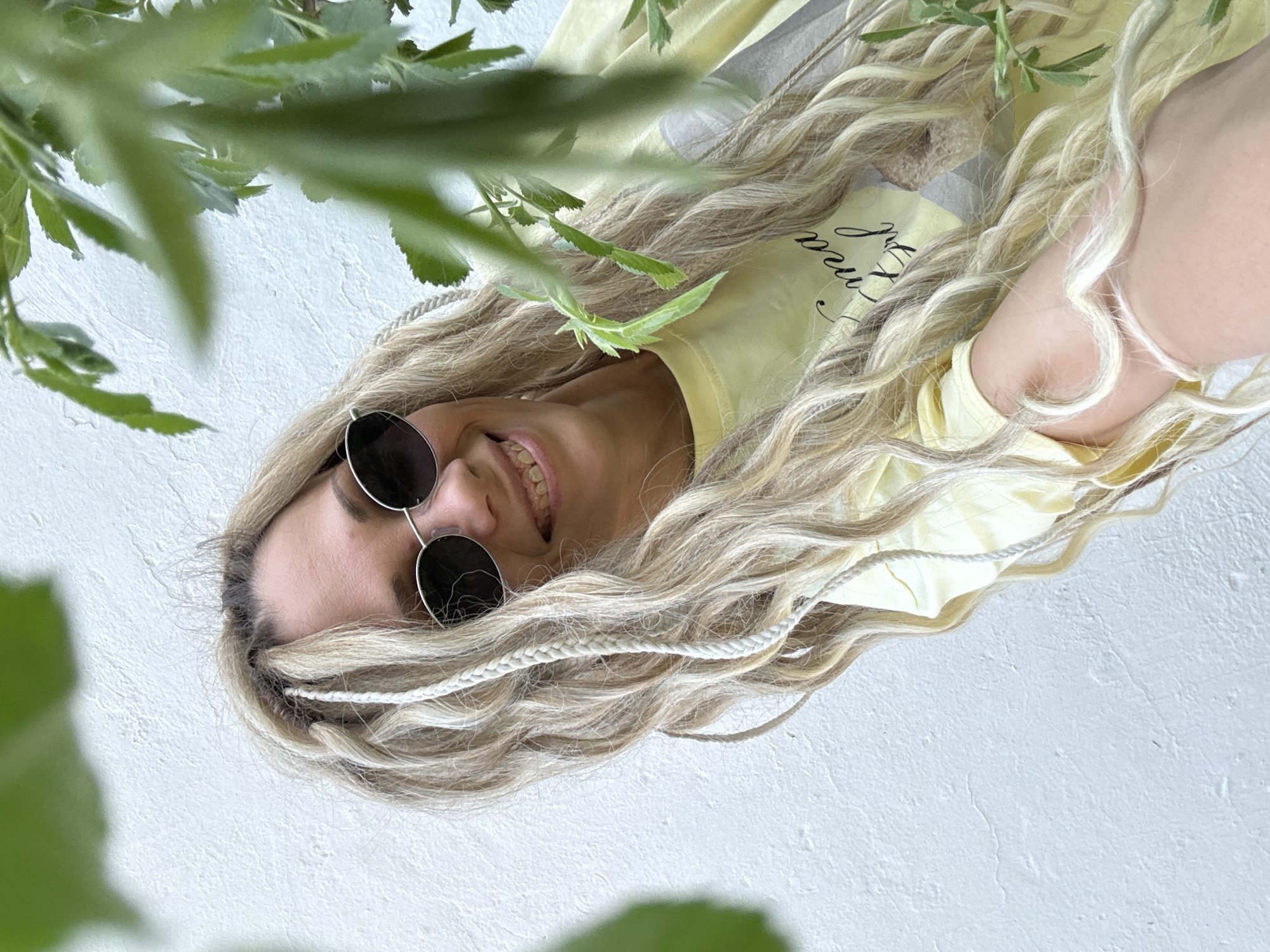A woman with long blonde hair with white braids, wearing round sunglasses and a yellow shirt, is smiling into the camera. The background is a textured white surface with green foliage partially obscuring the view. The image seems to be taken from a low angle, looking up at the woman. There's no verifiable information about the location being in Шебунино, Russia within the image context.