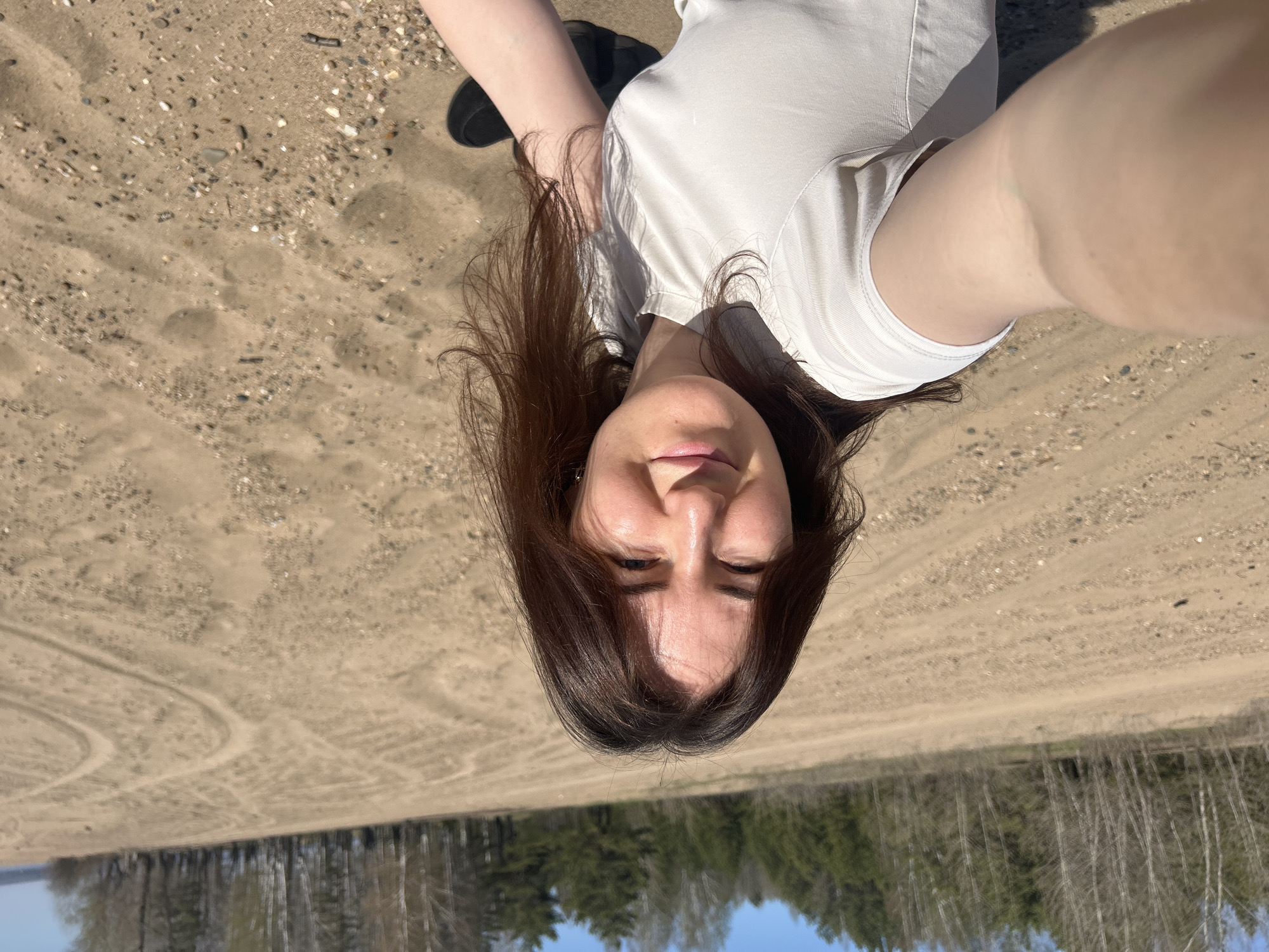 A woman is lying on her back in a sandy area. She has dark hair and is wearing a white shirt. The sand appears to have tire tracks. A forest of trees is visible in the background. The sky is blue. The location of the city of Noviy, Russia is not discernible from the image.