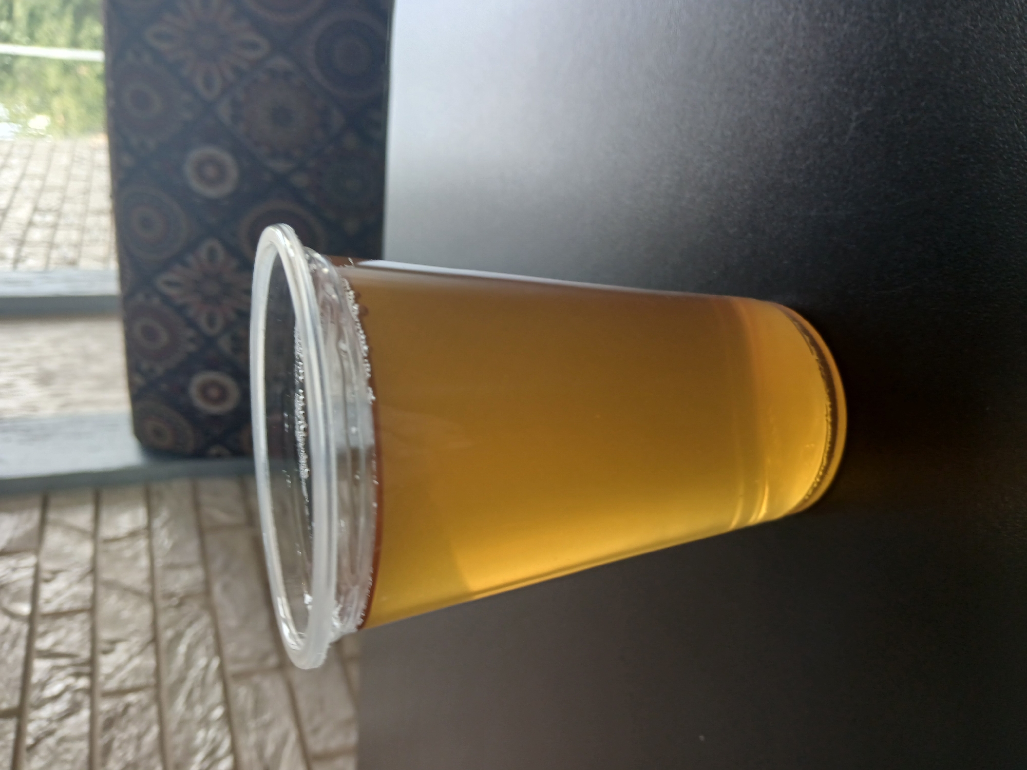 A close-up shot shows a clear plastic cup filled with a golden-amber liquid, possibly tea or juice, with a thin layer of foam on top. The cup is positioned on a dark, textured surface, likely a table. In the background, a brick wall with a textured pattern is visible, suggesting an indoor setting. Behind the brick wall, a patterned upholstery is present, with a hint of green foliage visible through a window or opening. The lighting is soft, casting subtle shadows. There is no discernible information about the location being in Moscow, Russia.
