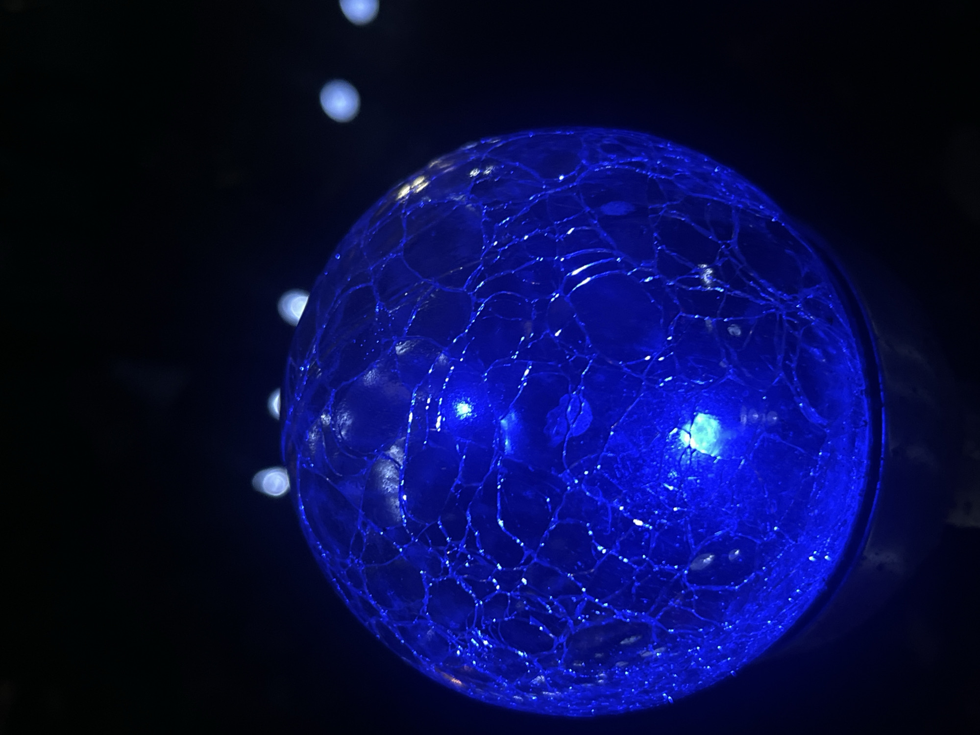 The image depicts a close-up, low-angle shot of a cracked, iridescent blue glass sphere illuminated from within. The sphere has a textured surface with a network of fine cracks that catch and refract the light. The background is dark and out of focus, with several bokeh-like light orbs visible in the upper right quadrant. The sphere is positioned on a dark, indistinct base, possibly a stand or fixture. The overall impression is one of abstract light and texture, with the sphere being the sole, clear subject. The image does not contain any identifiable elements that would place it in Branipole, Bulgaria, or any specific city. There are no people, actions, or interactions visible.