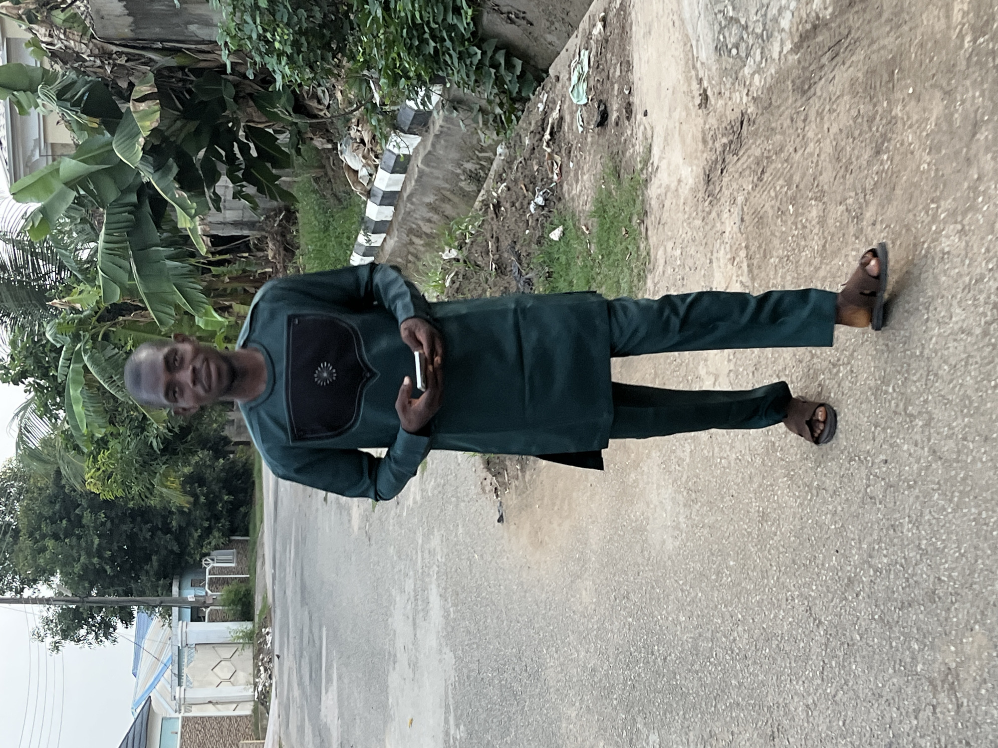 A man stands on a paved road, dressed in a dark green traditional African attire. He wears sandals and holds a small, rectangular device. Behind him, there is lush vegetation, a concrete structure with black and white stripes, and some scattered debris. In the background, residential buildings and trees are visible. This scene appears to be taking place in a residential area in Abuja, Nigeria.