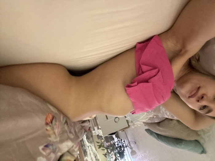 The image depicts a young adult female lying on her left side on a bed in what appears to be a bedroom. She faces the camera, her head resting on a pillow. Her right arm is positioned above her head. A bright pink garment is visible, pulled up to expose her midriff and hip, while the lower portion of her body, including her left thigh, is visible along the edge of the bed. The bed is covered with light-colored sheets. Behind her head, two pillows are present: one light blue/green and another light brown/beige. A patterned blanket, featuring feather motifs in various muted colors, is partially visible draped over the bed near her head. In the background, to the right, a white bedside table with a lamp is visible. Beyond the table, green foliage from indoor plants can be seen, along with what appears to be curtains and a light-colored wall with a subtle pattern. The lighting suggests an interior setting. There are no identifiable features within the image to determine a specific location within Saint Petersburg, Russia, or any other city.