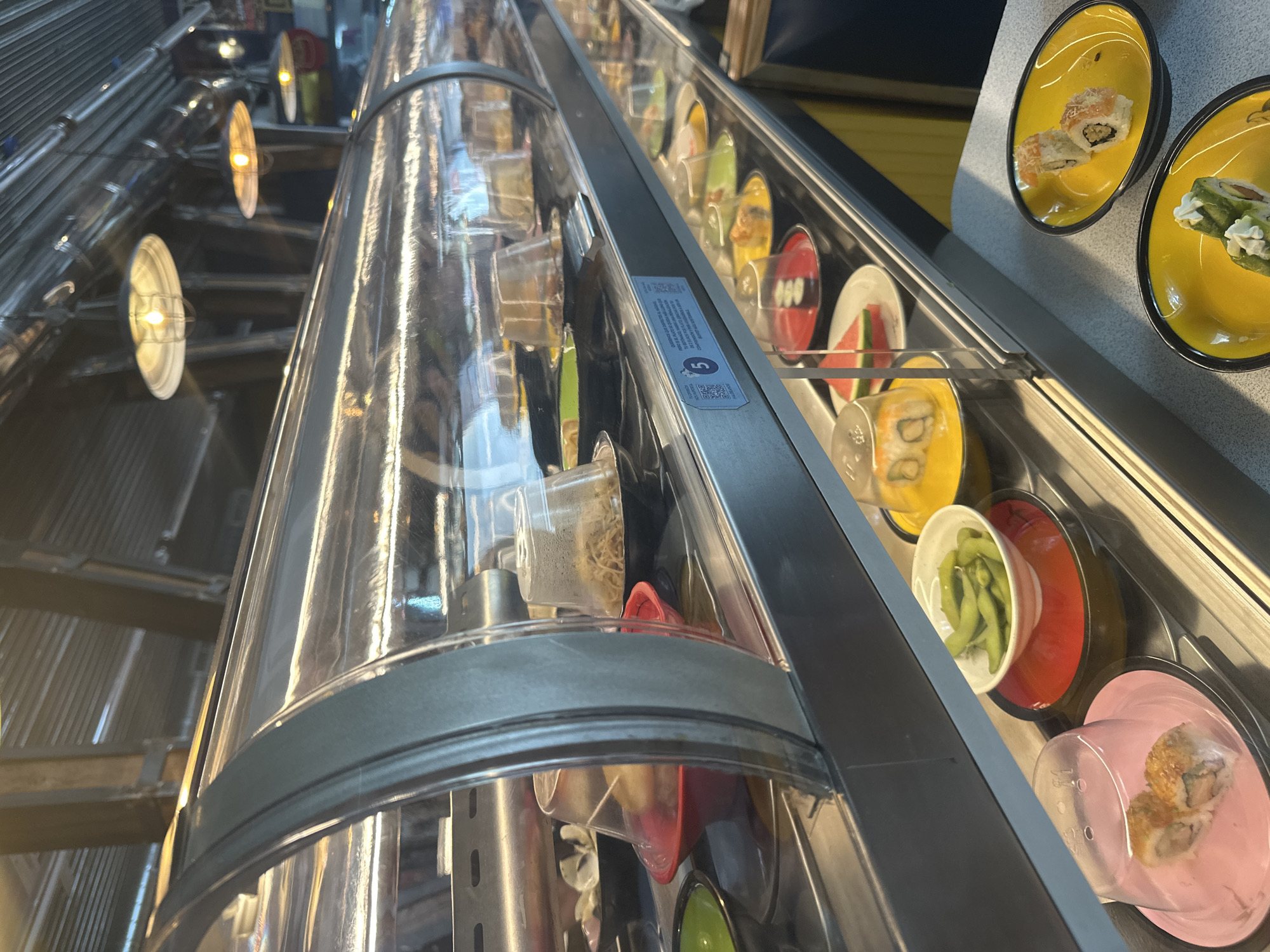 The image depicts a conveyor belt sushi restaurant, likely in Madrid, Spain, based on the prevalence of this type of restaurant in the city. The conveyor belt features a variety of dishes, including sushi rolls, edamame, and other small items, presented on colorful plates. The food is enclosed within a clear, cylindrical casing. The lighting is provided by industrial-style hanging lamps. The background suggests a modern interior with exposed metal structures.