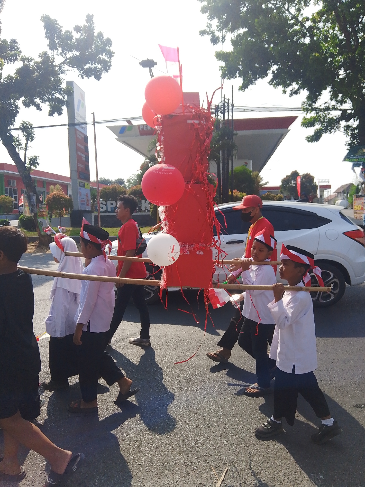 In the city of Rejosari, Indonesia, a daytime outdoor scene shows a parade or procession taking place on a paved street. The sun is bright, casting long shadows. In the foreground, several children are walking. Two boys in the center, dressed in white shirts and black pants, are carrying a large red, cylindrical structure on their shoulders using bamboo poles. They are also wearing red, white, and black headbands. To their left, a person in a white outfit, possibly a girl, also wears a similar headband and is being carried on the poles. Behind them, a young man in a red t-shirt and black pants walks, and further back, a man in a red cap and black mask also appears to be part of the group.
The red structure is decorated with red streamers and has red and white balloons attached to it. One white balloon is visible with text on it. In the background, a white car is driving alongside the procession. Across the street, there's a brightly colored building with a red roof, possibly a gas station, identified by a prominent vertical sign with blue and white elements. Trees with green foliage are visible in the upper parts of the image. The overall atmosphere suggests a festive or celebratory event.