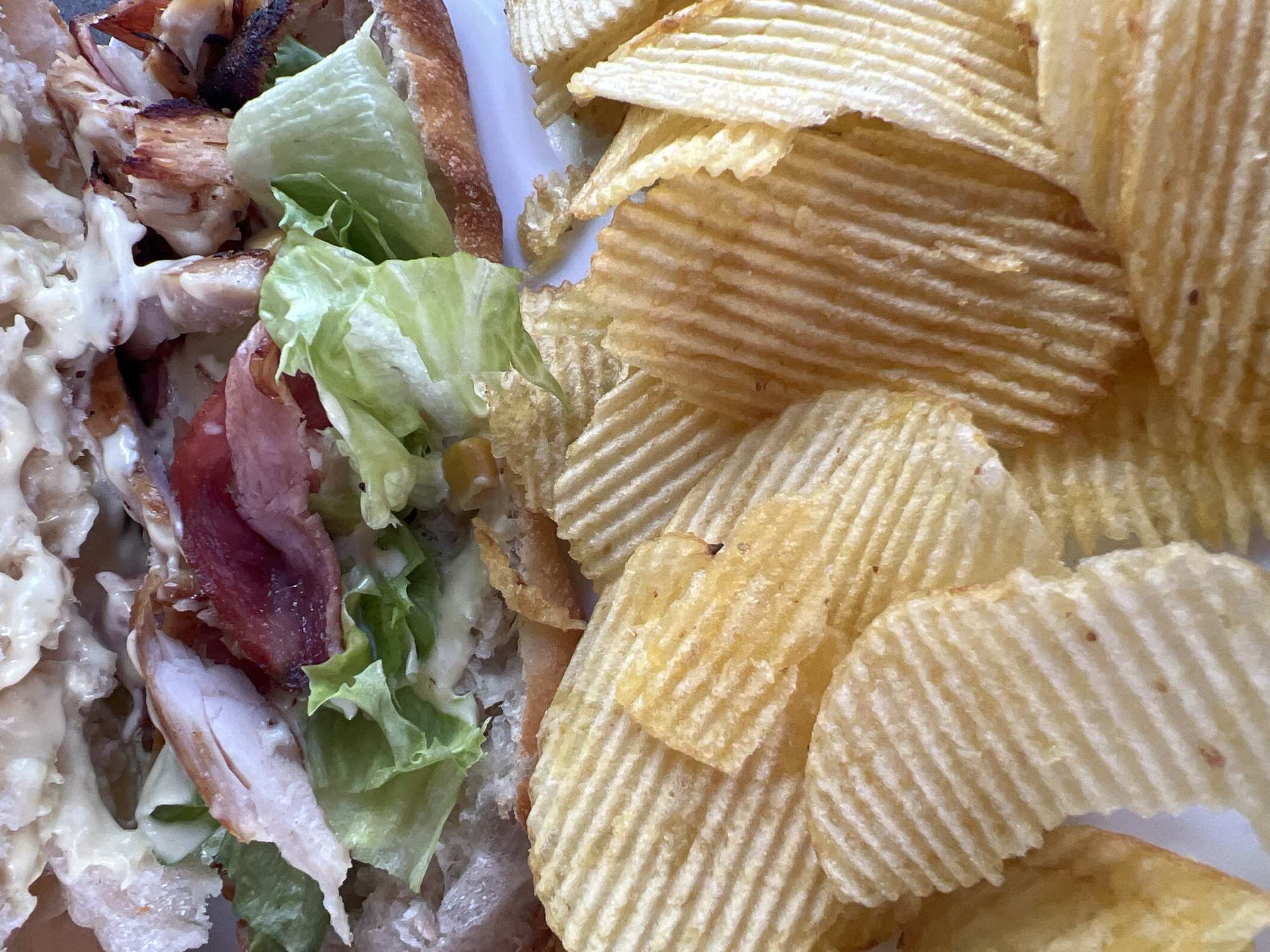 A close-up shot shows a sandwich filled with lettuce, chicken, and bacon, accompanied by a pile of ridged potato chips on a white plate. The lighting is natural, and the focus is on the food. The location is not discernible from the image.