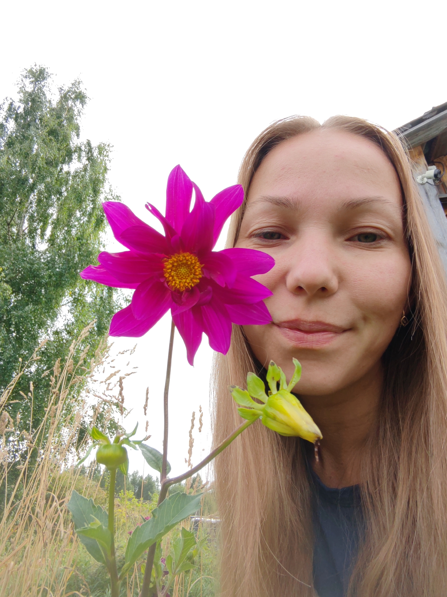 A close-up, low-angle selfie is captured outdoors, with a young woman's face filling the right side of the frame, her gaze directed towards the camera with a slight smile. Her long, blonde hair cascades down her shoulders. A vibrant pink dahlia flower is prominently positioned in the left half of the image, its petals unfurling. Below the dahlia, a green, unopened flower bud is visible. The background features dry, tall grass, green trees, and a glimpse of a wooden structure on the far right. The sky is overcast and white. The scene appears to be in a rural or garden setting.