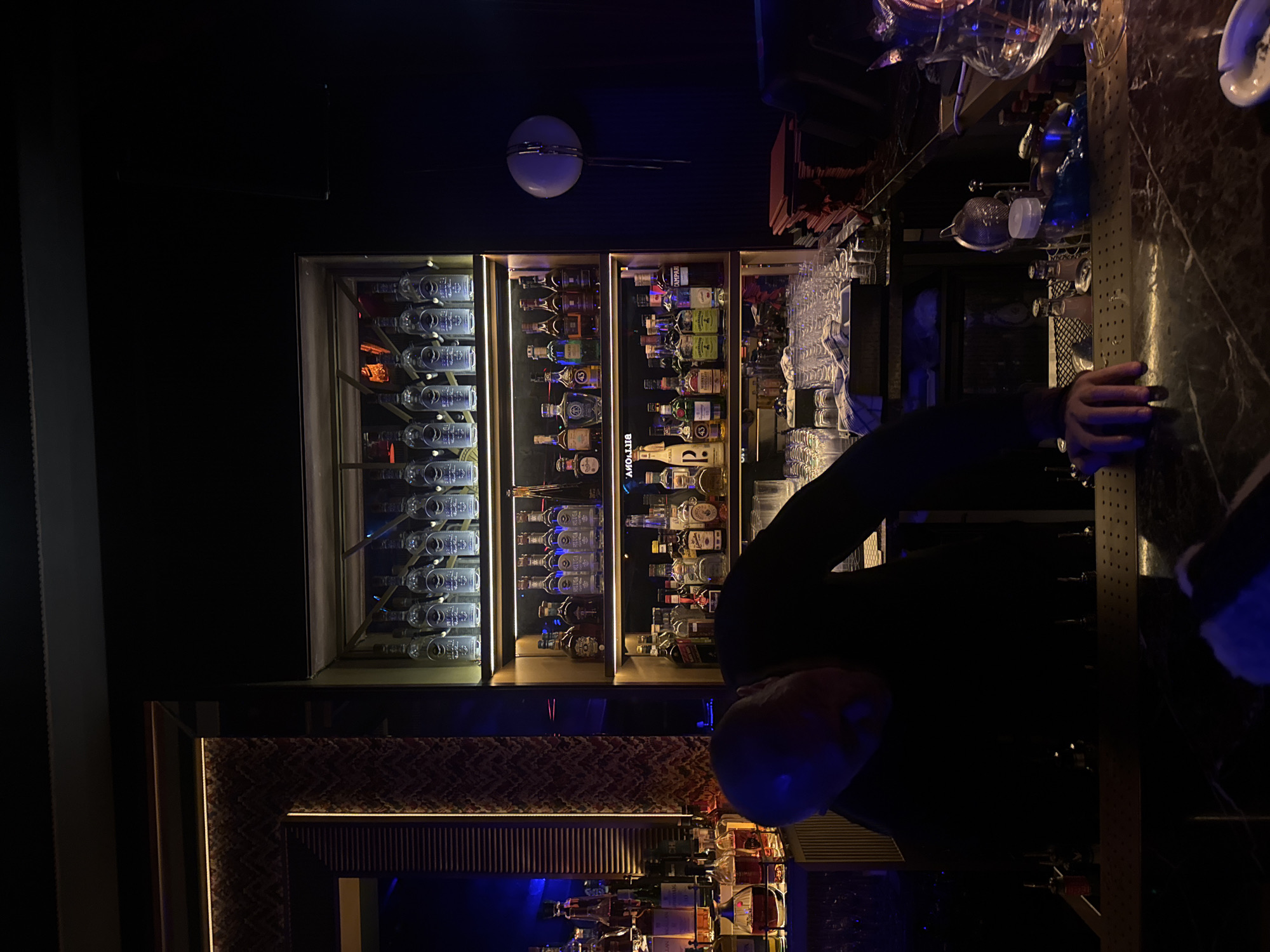 The image depicts the interior of a bar, possibly in Dubai, United Arab Emirates, based on the style and ambiance. Behind the bar is a well-lit liquor cabinet filled with bottles of alcohol and glassware. A person, likely a bartender, is partially visible in the foreground, positioned behind the bar. The bar itself has a textured surface. The lighting is dim and atmospheric, with a mix of warm and blue tones. A decorative element, perhaps part of the bar's design, is visible below the liquor cabinet. The overall setting suggests a sophisticated and modern establishment.