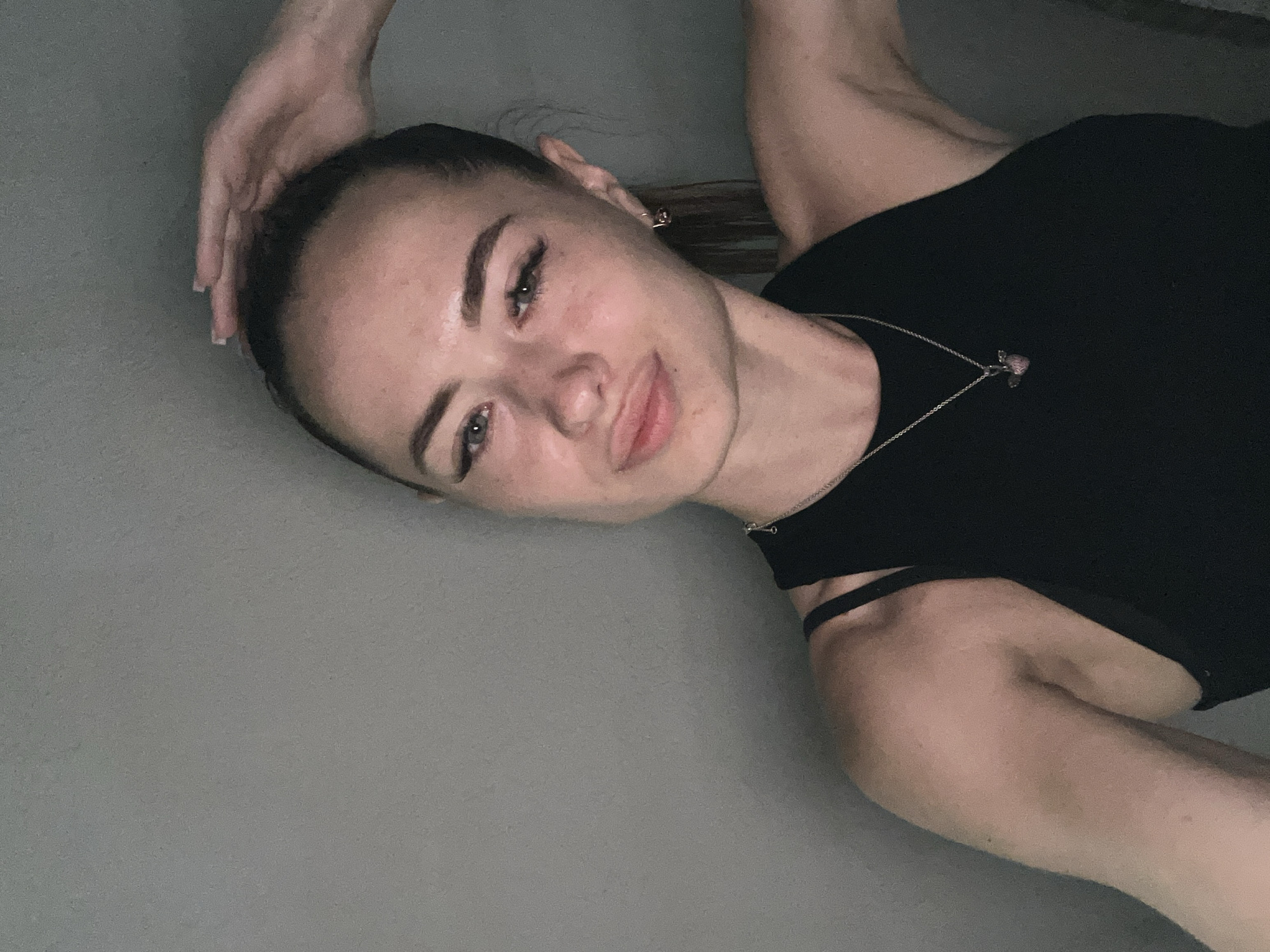 A young Caucasian woman with long, dark hair pulled back into a ponytail, is shown in a close-up, waist-up, indoor selfie. She is wearing a black, sleeveless tank top and a silver necklace with a pink pendant. Her right hand is resting on top of her head. The background is a plain, gray wall. She has fair skin with light freckles and light-colored eyes, and she is wearing makeup, including winged eyeliner and mascara.