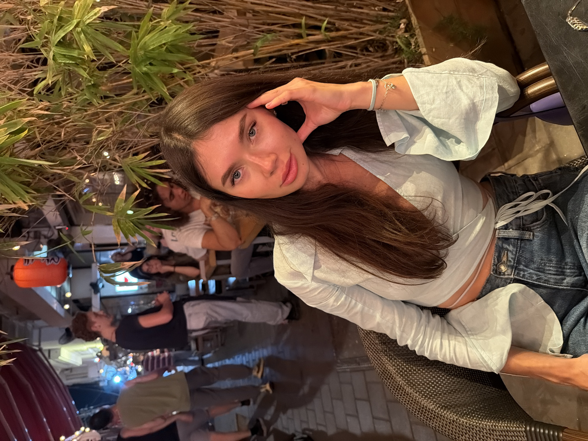 A woman with long, dark brown hair sits in a chair at an outdoor restaurant, with bamboo plants behind her and other diners visible in the background. She is wearing a light blue, open-fronted shirt with wide sleeves and blue jeans. She is looking at the camera with her right hand resting on her cheek. The location appears to be an alleyway in Tel-Aviv, Israel, suggested by the red awning and the general atmosphere of an outdoor dining area. The image is taken at night, with artificial lighting illuminating the scene.