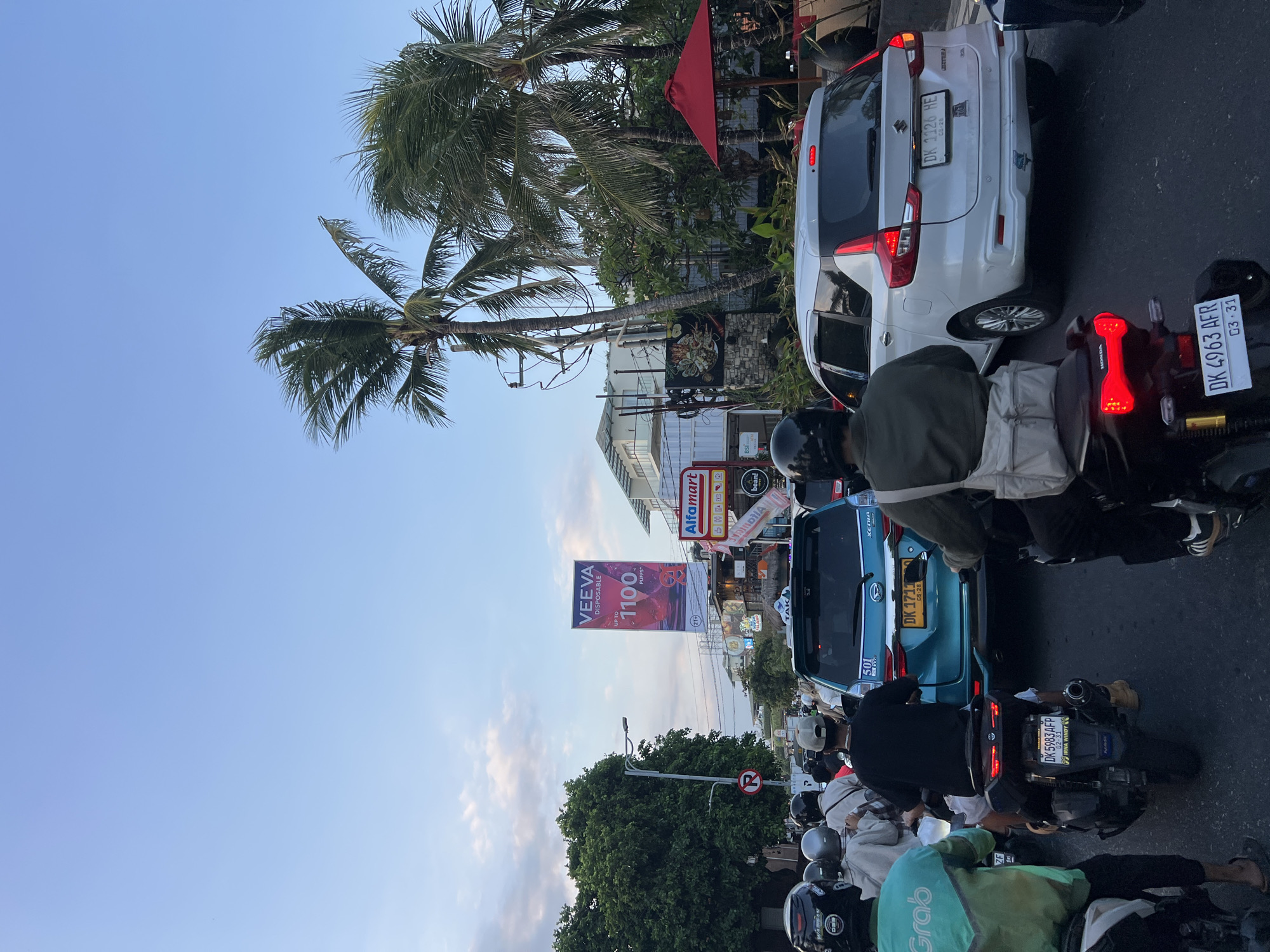 The image depicts a traffic jam on a street in Kuta, Bali, Indonesia. The sky is a pale blue with scattered clouds. Palm trees line the sides of the road, some of which appear to be swaying. Various vehicles, including motorcycles and cars, are stopped in traffic. Many of the motorcyclists are wearing helmets. A turquoise car with a license plate starting with "DK" is in the center of the frame. A white SUV with a license plate also starting with "DK" is to the right of the turquoise car. Billboards and signs are visible, including one for "Veeva Disposable" and another for "Alfamart." The overall impression is one of a busy urban environment during daylight hours, likely late afternoon or early evening, given the lighting.