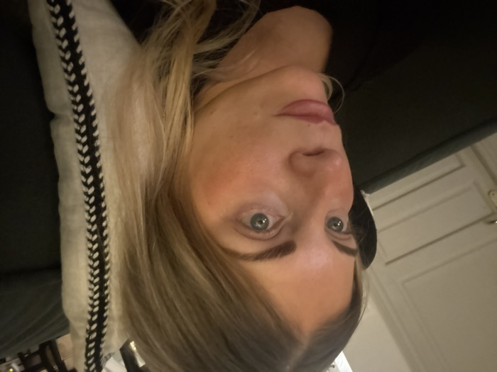 The image captures an adult female from the chest up, positioned indoors within the city of Rueil-Malmaison, France. She has light blonde hair, light blue-grey eyes, and defined eyebrows. Her complexion is fair with a visible flush on her cheeks, and her lips are full, colored with a light rosy-brown tone. She wears a dark-colored, likely black, garment on her upper body. Her gaze is directed off-camera towards the left of the frame, slightly upwards. She appears to be reclining against a dark piece of furniture, with a light-colored decorative pillow, featuring a prominent black braided vertical pattern, positioned to her right. A light-colored, panelled door with a metallic handle is partially visible in the upper left background. The overall lighting suggests an indoor, possibly evening, setting.