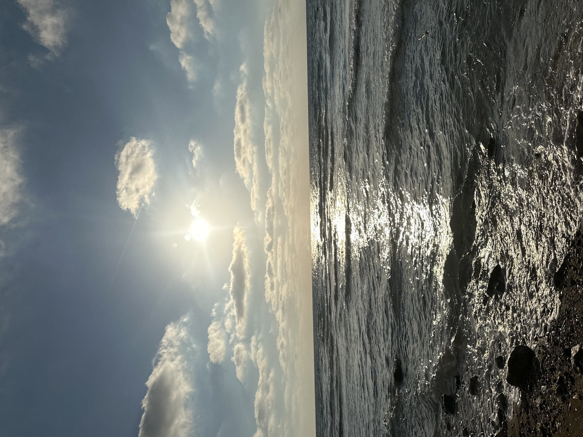 A serene daytime view captures the Mediterranean Sea at a beach in Haifa, Israel. The sun, positioned high in the bright blue sky, shines intensely, casting a brilliant reflection across the rippling water. Scattered white clouds dot the sky, some illuminated by the sun's rays. Gentle waves lap at the pebbly shore, where dark stones and wet sand are visible. The scene is devoid of any people or visible wildlife, creating a tranquil and natural landscape.