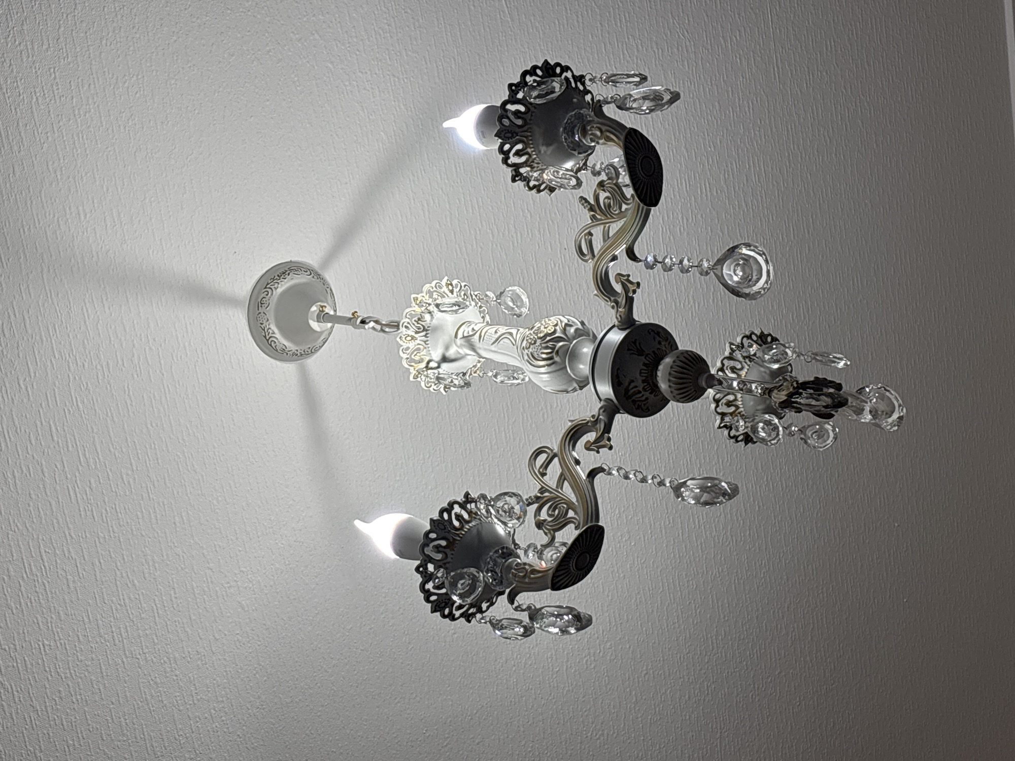 The image displays a close-up, low-angle view of a white and metallic chandelier hanging from a textured white ceiling. The chandelier has multiple arms, each adorned with decorative metalwork and crystal pendants. Three faux candle-shaped light bulbs are visible, emitting a soft glow. The overall aesthetic suggests an interior setting. There is no visible indication of the city of Getahovit, Armenia, or any other specific geographical location. The scene is static and devoid of any activity or people.
