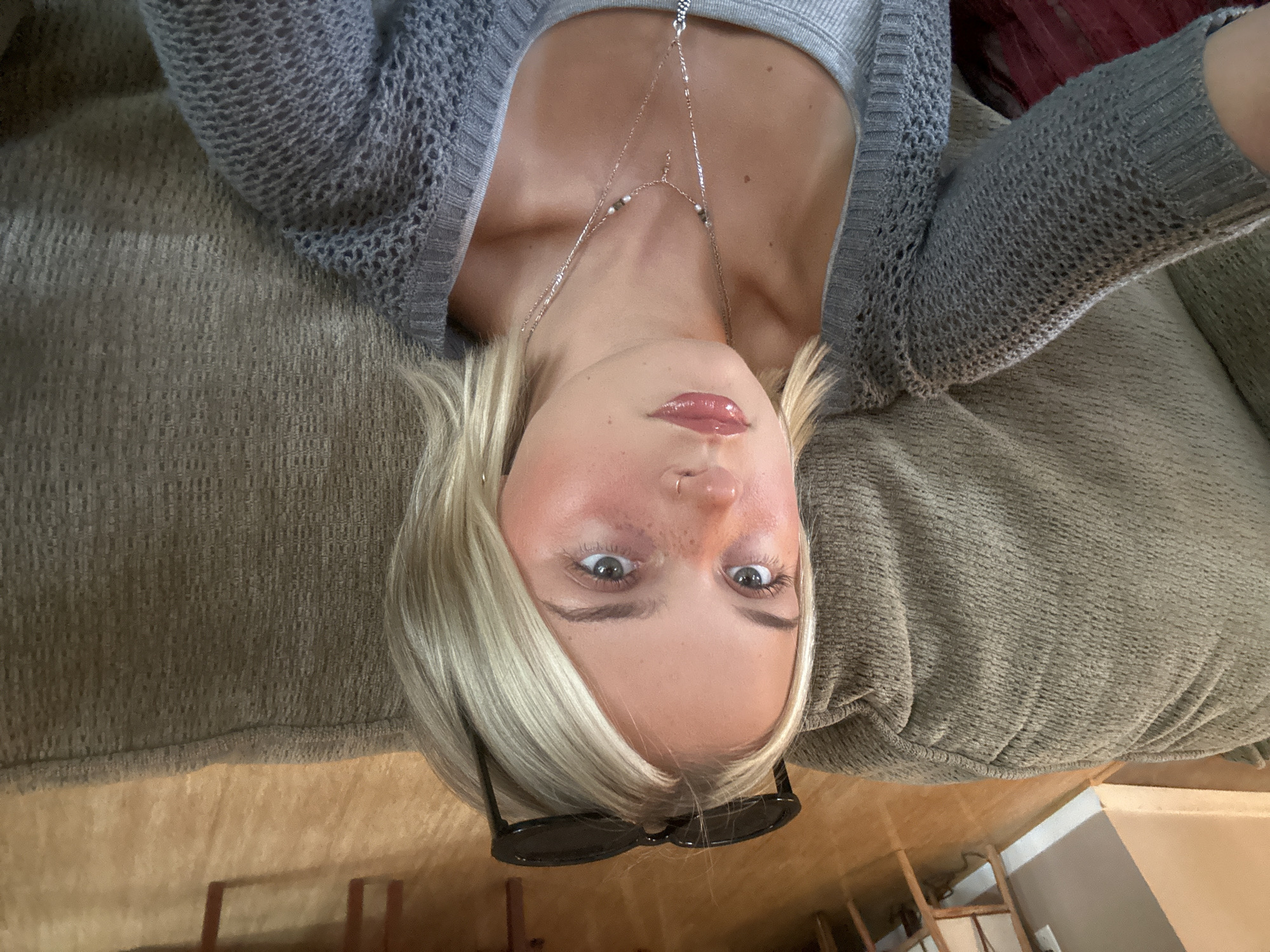 A young woman with blonde hair and sunglasses perched on her head is looking up and to the left. She is wearing a gray knitted cardigan over a gray tank top and multiple layered silver necklaces. She has light makeup on, including blush, mascara, and lip gloss, and a small nose ring. She is seated on a textured, olive-green sofa. The background suggests an indoor setting with a wooden floor and furniture visible. The location is not specified as Whistler, Canada.