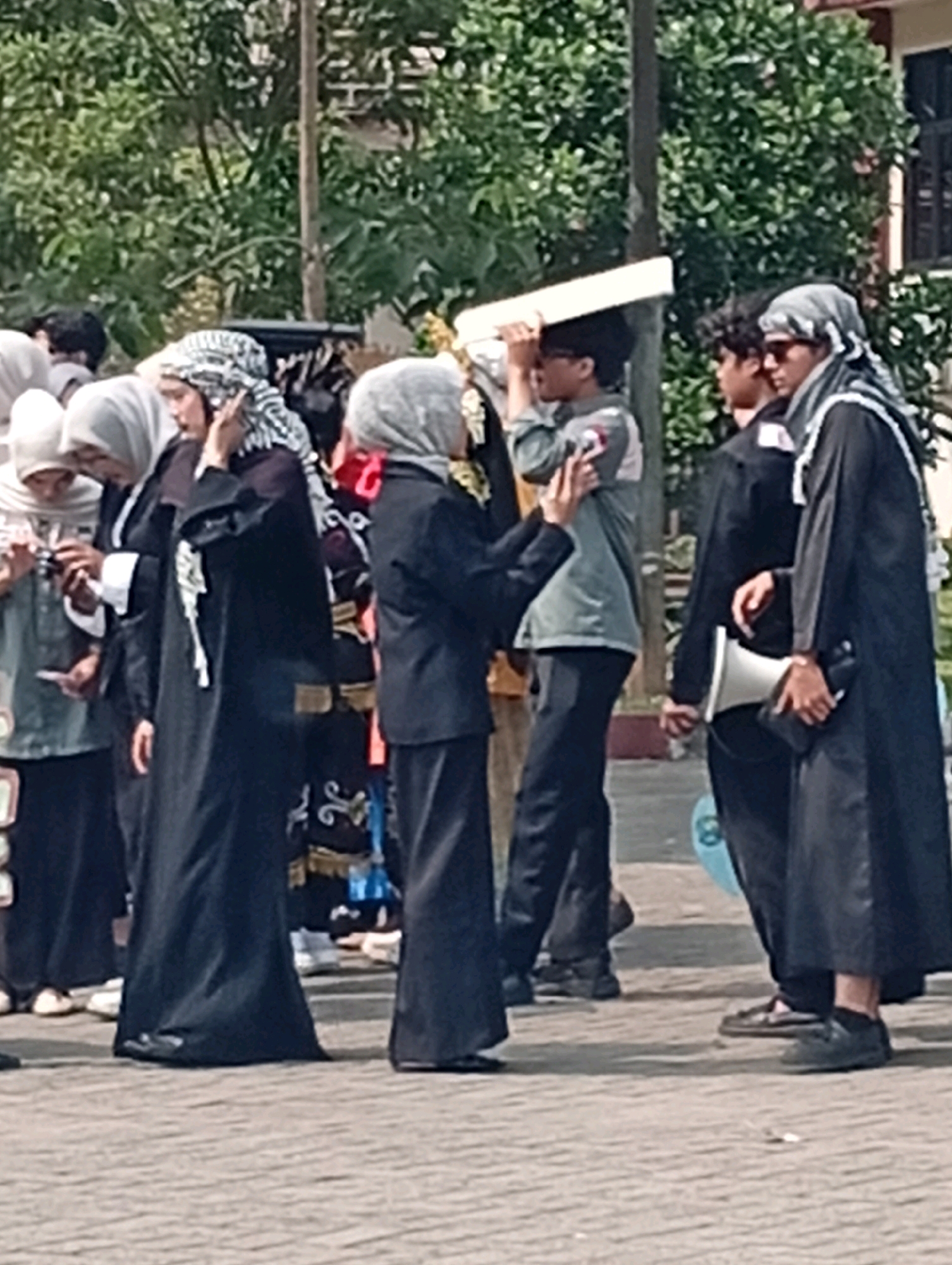 In Kelapa Dua, Indonesia, a group of individuals stands on a paved outdoor area, possibly a school campus. The primary subjects are a group of four people, three wearing traditional Islamic attire. On the far left, a person in a white hijab and a black abaya is partially visible. To their right, another person in a black abaya and a checkered headscarf is holding a smartphone, appearing to take a picture or record a video. Next to them, a person in a grey uniform and black pants is holding a flat white object above their head, and a person in black abaya and a grey headscarf stands to their right, holding a white megaphone. The background consists of trees and a building with windows. The scene suggests a public gathering or event.