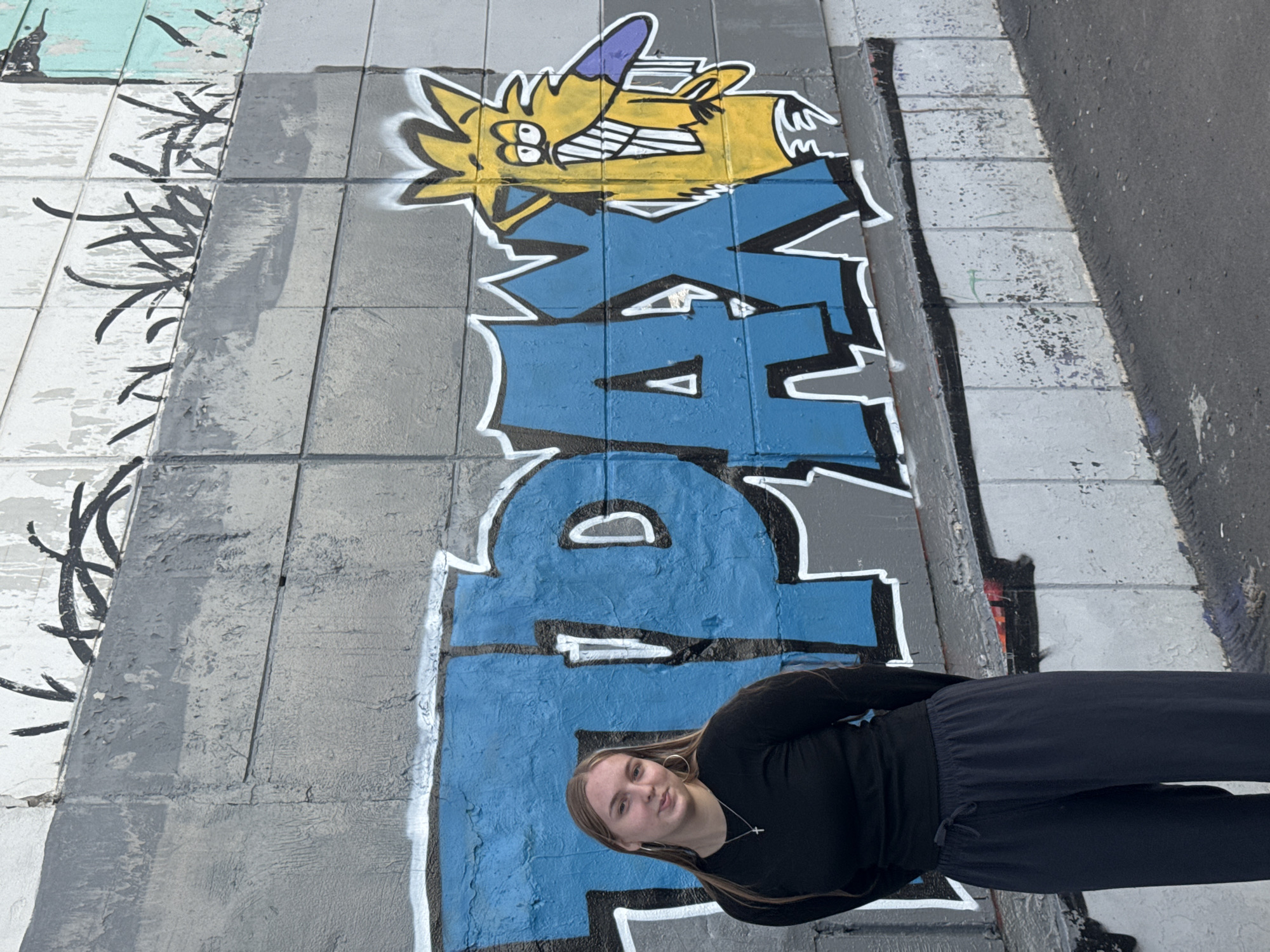 A young Caucasian woman with long blonde hair and hoop earrings stands in front of a graffiti-covered wall. She is wearing a black long-sleeved shirt and dark pants. The graffiti features large blue letters with white outlines and a yellow cartoon character with sharp teeth and a purple nose. The wall is made of concrete blocks. The scene appears to be an outdoor urban environment. This specific graffiti, featuring the letters "TPAX" and a caricature resembling Norbert from "The Angry Beavers," is located on a wall near a pedestrian bridge on the embankment of the Kama River in Perm, Russia. The area is known for its street art and is a popular spot for locals and tourists.