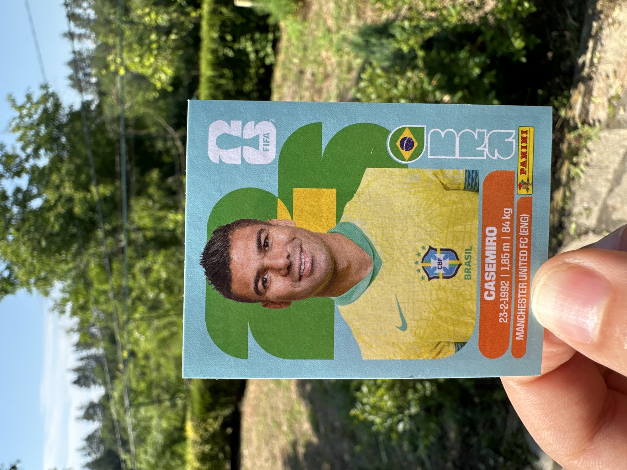 This media file shows a close-up, slightly angled view of a person's hand holding a collectible soccer sticker. The sticker features a portrait of a soccer player, identified by the text as "CASEMIRO," wearing a yellow Brazilian national team jersey with the "CBF" logo. The player's name is prominently displayed, along with his birthdate (23-2-1992), height (1.85 m), weight (84 kg), and current club (MANCHESTER UNITED FC (ENG)). The sticker also includes the "FIFA" and "PANINI" logos, indicating its origin and licensing.
The background of the image is blurred, but it appears to be an outdoor setting with green foliage, suggesting a daytime scene with natural light. The presence of trees and grass hints at a park or garden. The sunlight appears bright, casting shadows and illuminating the scene. The hand holding the sticker is in sharp focus, with visible skin texture and fingernails. The sticker itself is colorful, with a light blue background and vibrant graphics. The overall impression is that of someone showcasing a prized soccer collectible outdoors.
